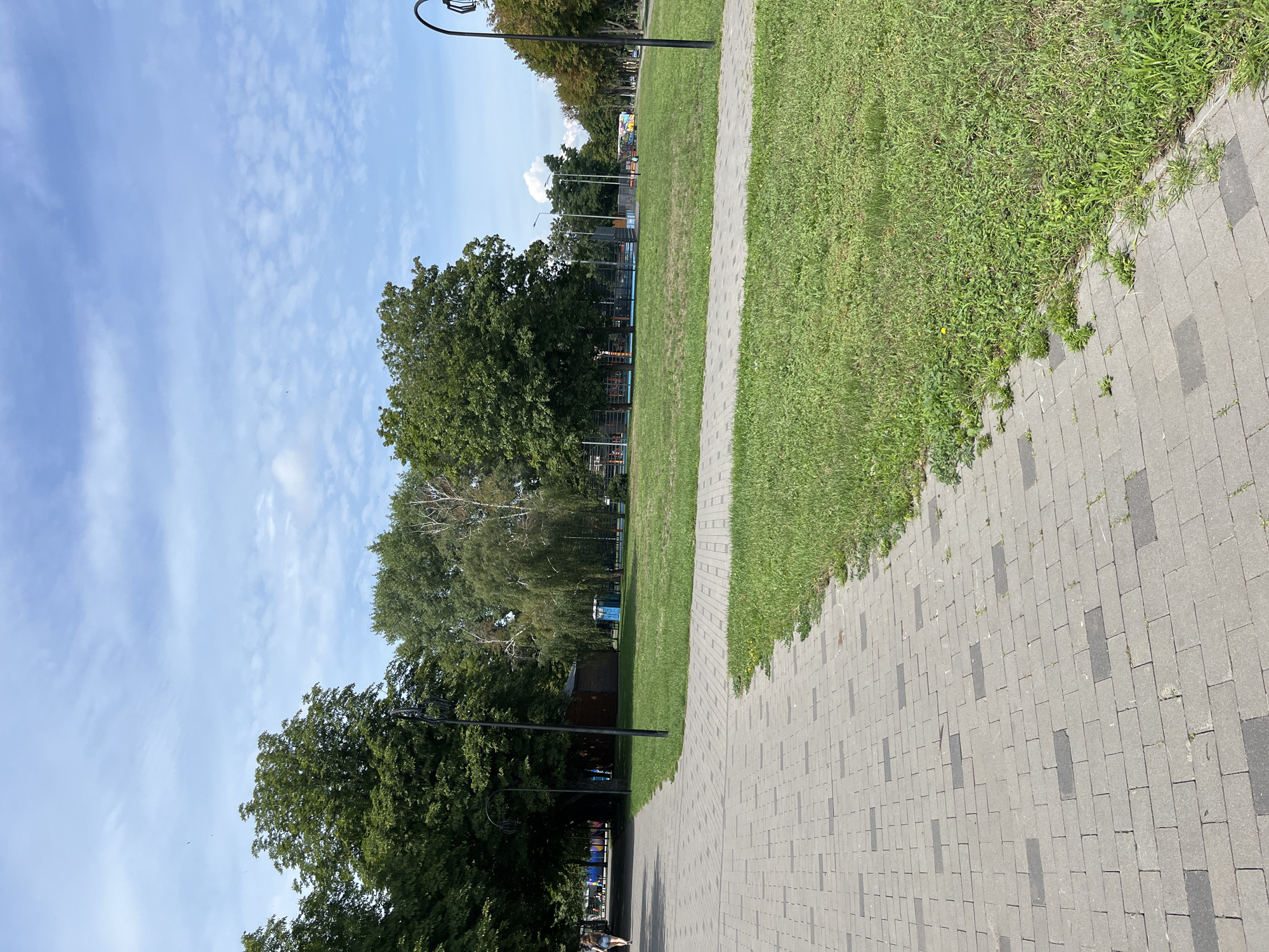 The image depicts a wide paved walkway in a park on a sunny day. The walkway is constructed with grey pavers, laid in a pattern, and is bordered by a well-maintained green lawn. Tall trees with lush green foliage line the edges of the park. In the background, there are glimpses of sports courts with blue flooring and orange markings, suggesting recreational facilities. A few people are visible in the distance, one walking on the path and others near the courts. The sky is blue with scattered white clouds. The scene appears to be a public park within the city of Kharkiv, Ukraine, likely a place for recreation and relaxation.