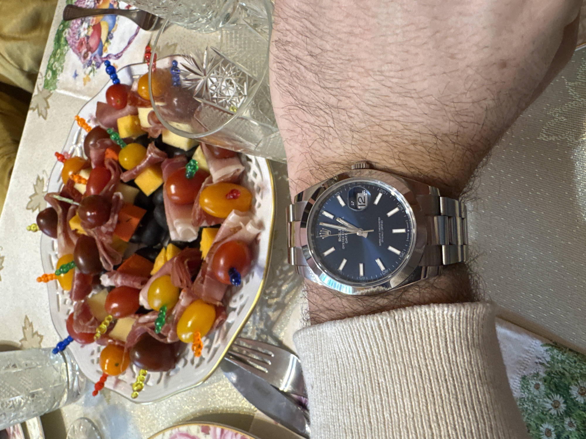 A person is wearing a silver Rolex Oyster Perpetual Datejust watch with a blue dial on their left wrist. The person is wearing a cream-colored, knitted sweater. To the left of the arm and watch is a small, decorative plate of hors d'oeuvres. The hors d'oeuvres consist of cherry tomatoes, cheese cubes, olives, and thin slices of ham or prosciutto, all skewered with colorful plastic toothpicks. A cut-crystal glass sits partially on the plate, obscuring some of the hors d'oeuvres. Silver cutlery, including a knife and fork, is visible near the plate. The tablecloth has a floral print with leafy accents. The location is unspecified and there is no evidence that the location is Plovdiv, Bulgaria.