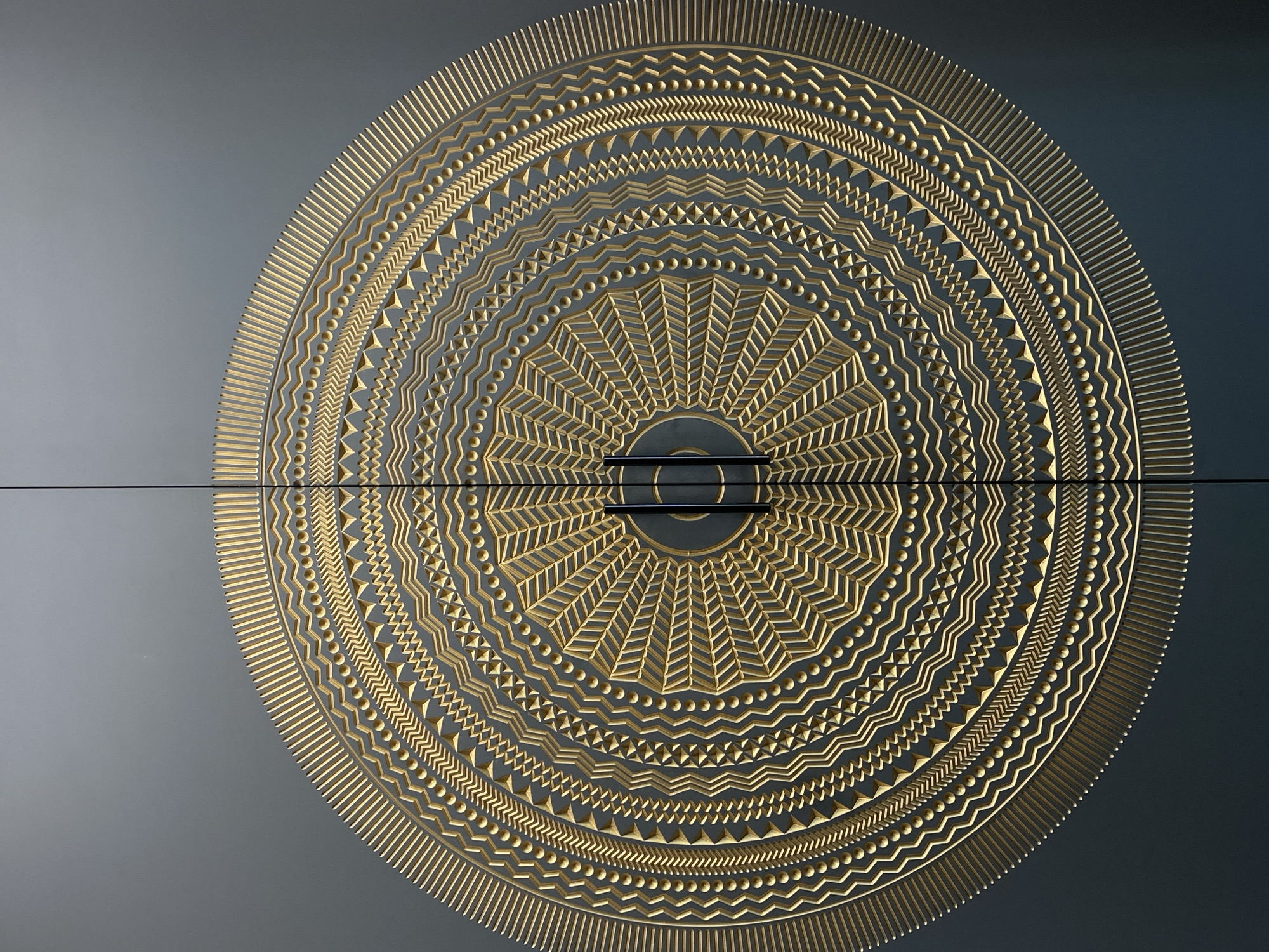 A close-up shot showcases a circular, decorative design in a gold tone against a grey-blue background. The design is centered and appears to span across two horizontal panels with a thin black line separating them. The design consists of concentric rings, each featuring unique geometric patterns. The innermost ring displays a radiating, fan-like pattern of lines, followed by rings featuring chevrons, lines of spheres, zigzag patterns, and radial lines. The image does not depict people, subjects, or actions. The environment is limited to the surface showcasing the design. The location in Votkinsk, Russia is unknown.
