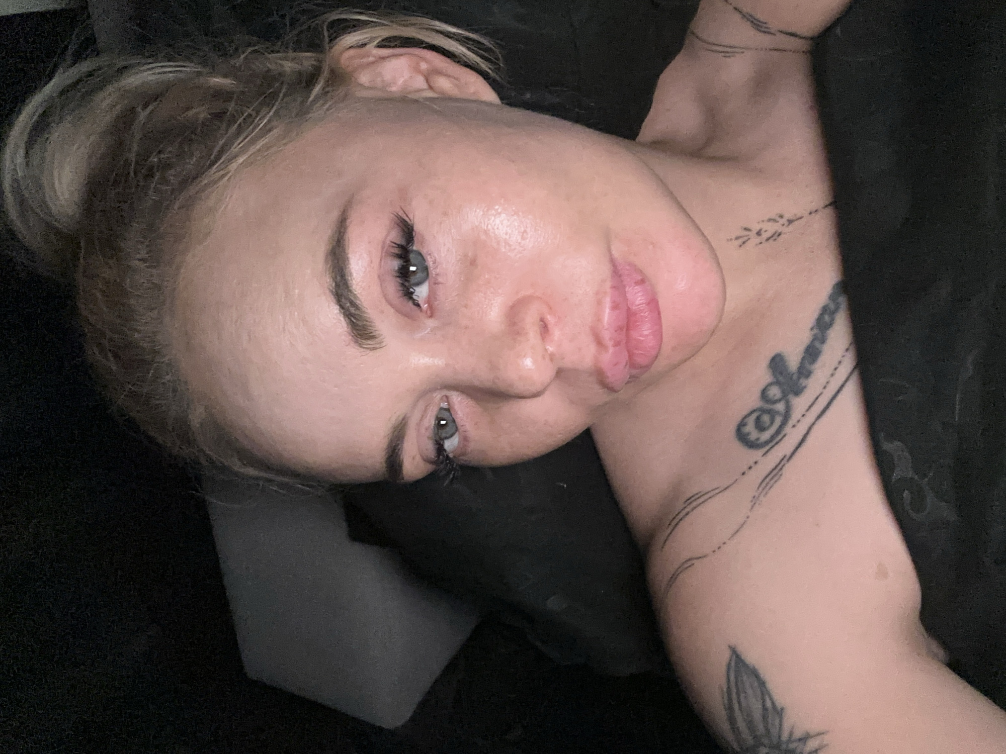 A close-up, dimly lit portrait captures an adult female in an indoor setting, location Unknown, Unknown. The subject has light skin, blonde hair pulled into a high, messy bun, and blue-grey eyes. Her gaze is directed towards the camera, and her expression features full, slightly parted lips in a pout. Her face appears somewhat shiny, with visible pores, light freckles, and prominent dark eyelashes, possibly extensions.
Several tattoos are visible on her upper torso. On her left shoulder (viewer's left), a partial outline of a dark wing design is present. On her right collarbone and upper chest area (viewer's right), script lettering, possibly "Aura," is visible with extending decorative lines. Below the center of her collarbone, a vertical tattoo resembling a stylized wheat stalk or feather is inked. Additional thin, parallel lines are visible on her outer right shoulder.
She is partially reclined, possibly in a bed, with a dark, indistinct fabric covering her lower chest and shoulders. The background is uniformly dark and out of focus, indicating a dimly lit interior space with soft furnishings, possibly a pillow or headboard behind her. The lighting appears to be soft and ambient, illuminating her face from the front.
