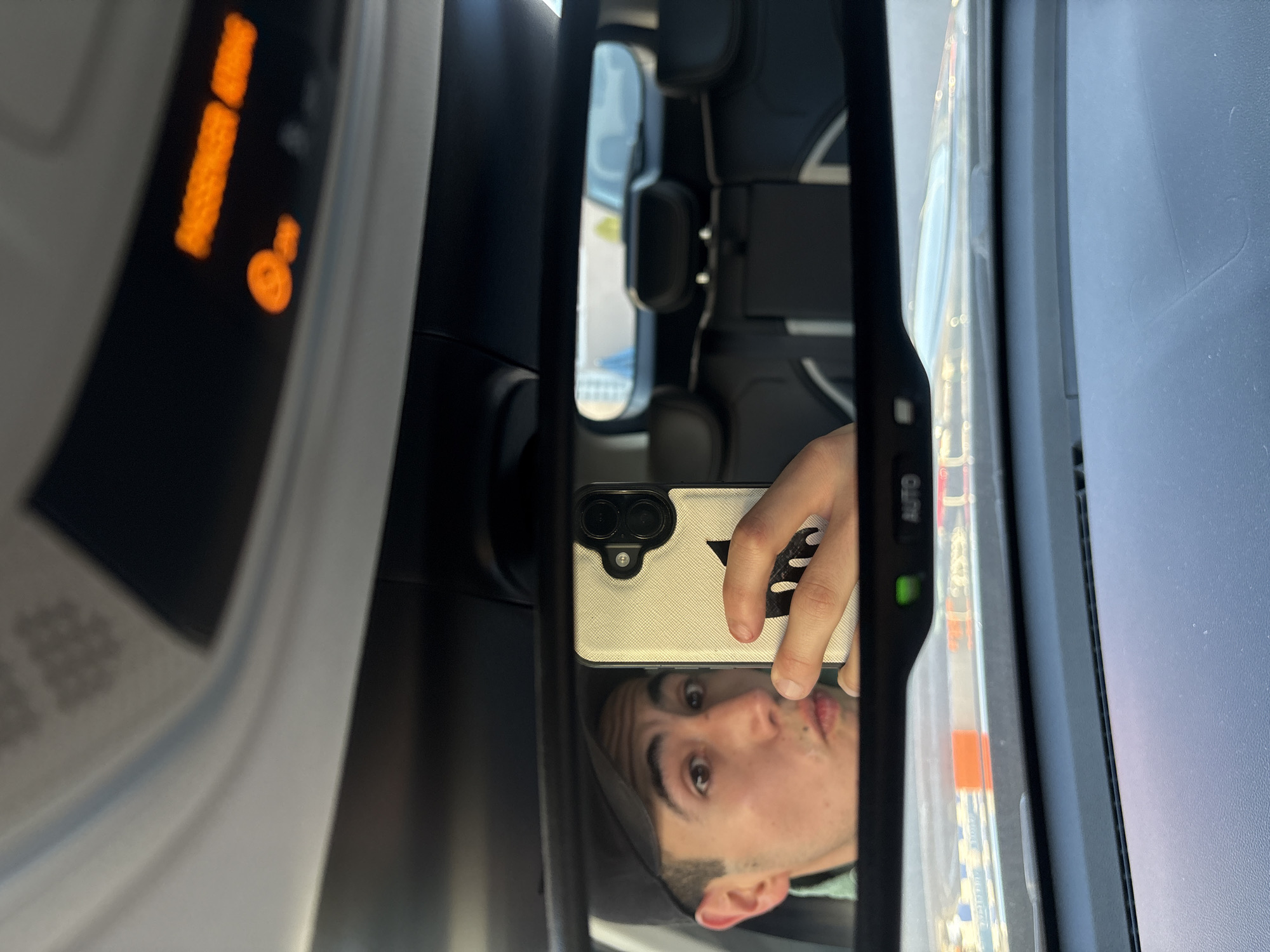 The image captures a reflection in a car's rearview mirror, taken from the driver's or passenger's seat. The primary subject is a young man, whose face is reflected in the mirror, looking directly into the camera of the smartphone he is holding. He has dark hair and a relaxed, slightly puckered expression.
The man is holding a smartphone with a light-colored, textured case that features a black triangular design and a prominent black dual-lens camera module. His hand and part of his arm are also visible in the reflection.
The rearview mirror itself has a black frame, and on its right edge, the word "AUTO" is visible in white letters next to a small green indicator light. Above the mirror, the car's overhead console is partially visible, showing illuminated orange digital segments and an icon, possibly part of a display or control panel. Through the mirror and in the background, elements of the car's dark interior, such as headrests and seats, are faintly discernible. Faint, blurry reflections of the exterior environment suggest it is daytime. The scene is set inside a vehicle in San Bernardino Tlaxcalancingo, Mexico.