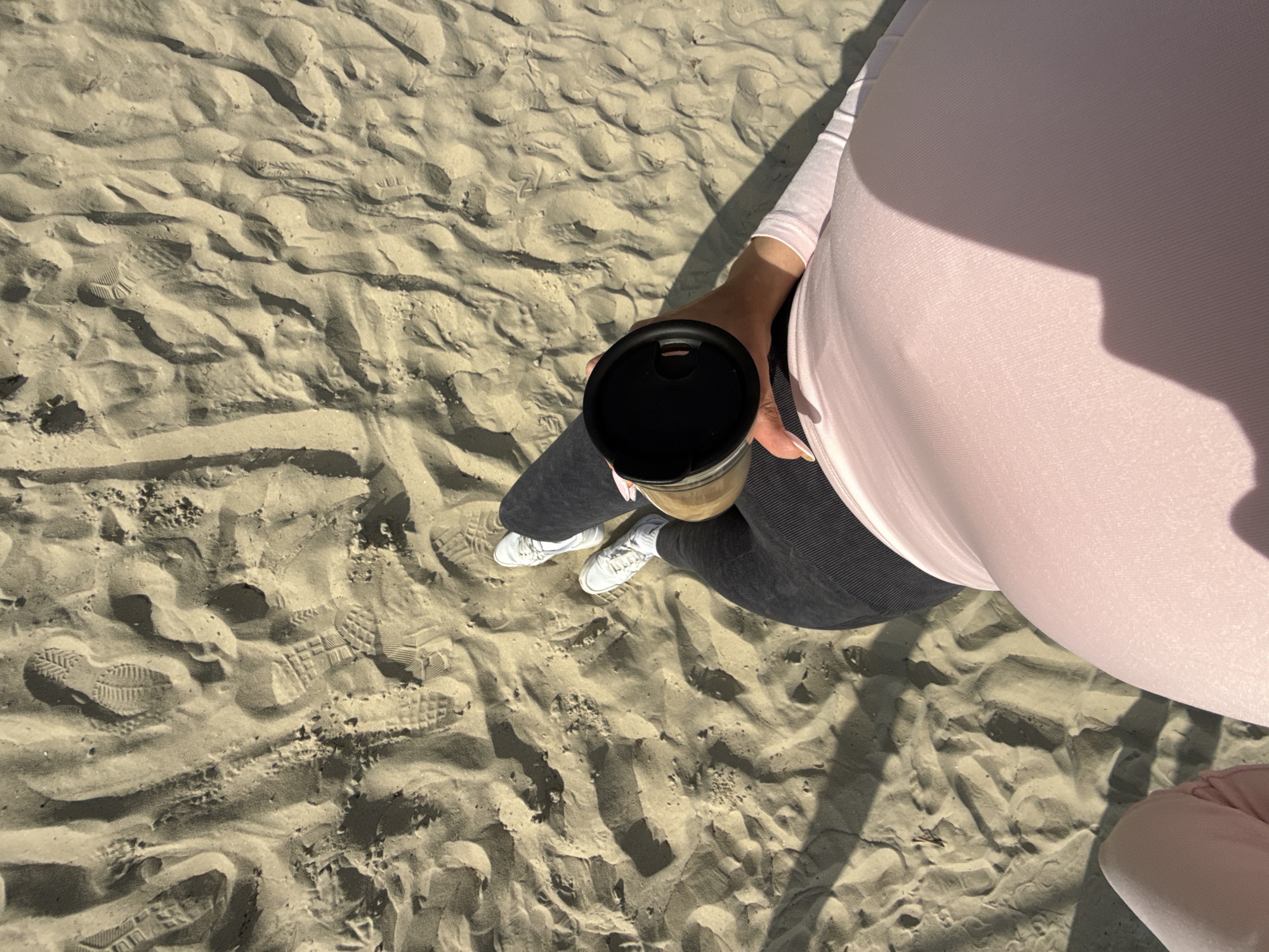 A high-angle shot captures a pregnant person standing on sand, possibly at a beach. The person is wearing a light pink long-sleeved shirt and dark grey leggings. They are holding a travel mug with a black lid. The person is wearing white sneakers. The sand shows numerous footprints and ripples. Given the location provided, the scene might be on a beach in or near the city of Nesebar, Bulgaria.