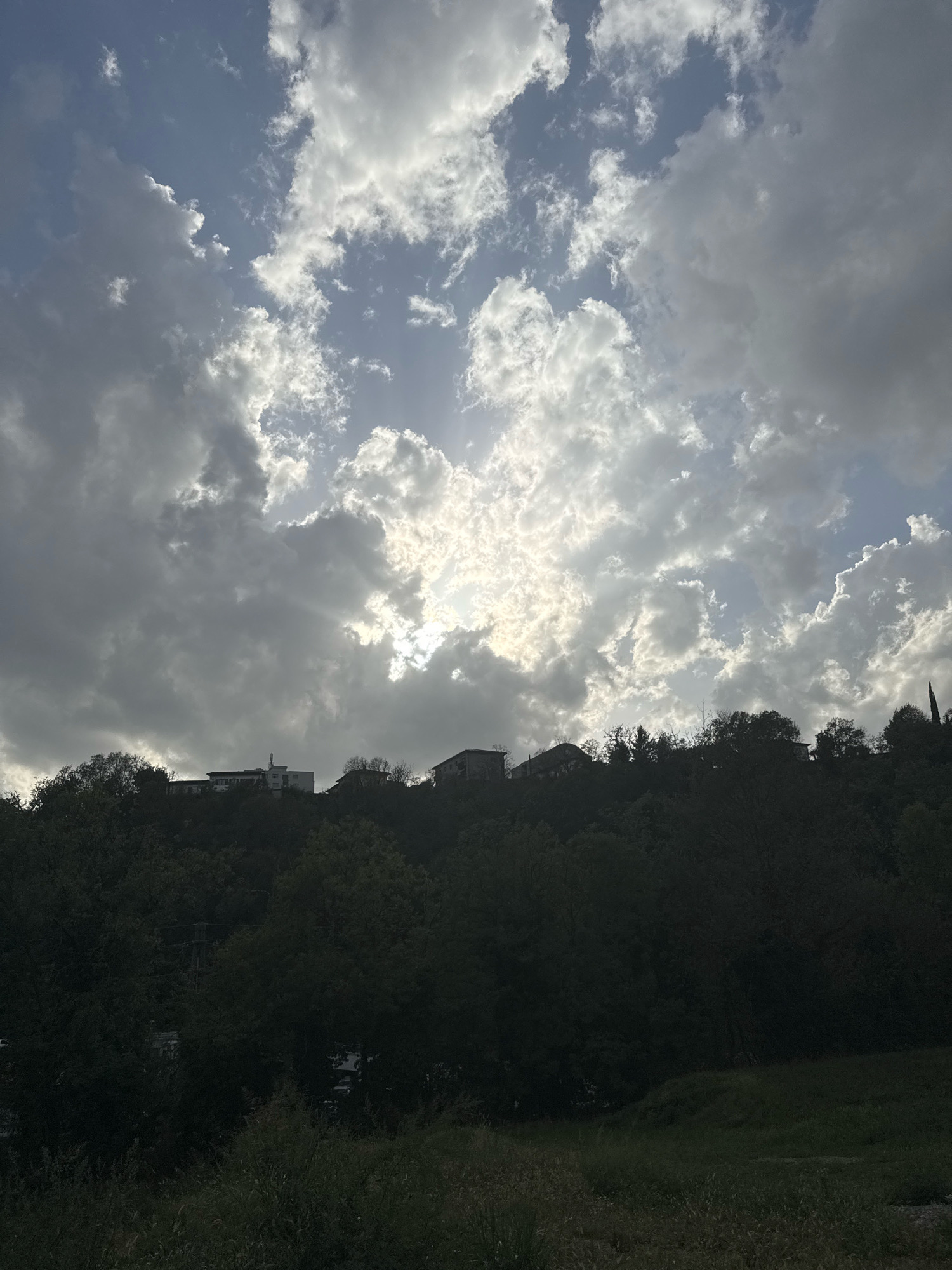 A dramatic sky fills the upper portion of the image, with a mix of dark, heavy clouds and bright, sunlit patches. Sun rays pierce through the clouds, creating a luminous effect. Below the sky is a silhouette of a tree-covered hillside. Several buildings are visible atop the hill, appearing as white or light-colored structures against the darker foliage. The foreground is a dark, grassy area. The scene appears to be a landscape view of a hillside with structures in Morbio Inferiore, Switzerland, under a dynamic sky.