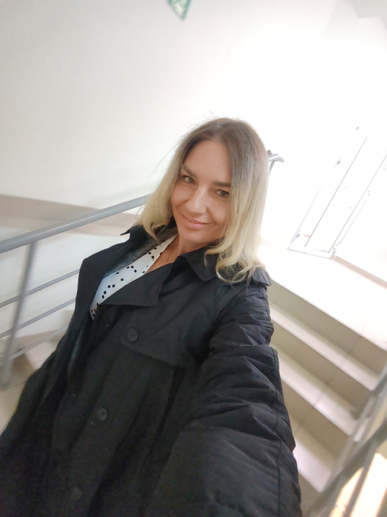 The image is a selfie of a woman taken indoors, likely within a building in Petropavlovsk-Kamchatsky, Russia. The woman, with shoulder-length blonde hair, is smiling and looking directly at the camera. She is wearing a black trench coat or jacket over a white top with black polka dots, which is partially visible. Her left arm is extended towards the camera, indicating she is holding the device herself.
The background consists of light-colored walls and a ceiling. To the left, a grey metal handrail is visible, suggesting a staircase. On the right side of the frame, a brightly illuminated area shows more stairs and what appears to be a window or a strong overhead light source, creating a significant amount of glare. This bright lighting indicates it is likely daytime. In the upper left corner of the image, a faint, indistinct light green symbol is partially visible on the ceiling, which could be an exit sign or another type of informational marker. The overall impression is one of a casual, self-taken portrait in a public or semi-public indoor setting.