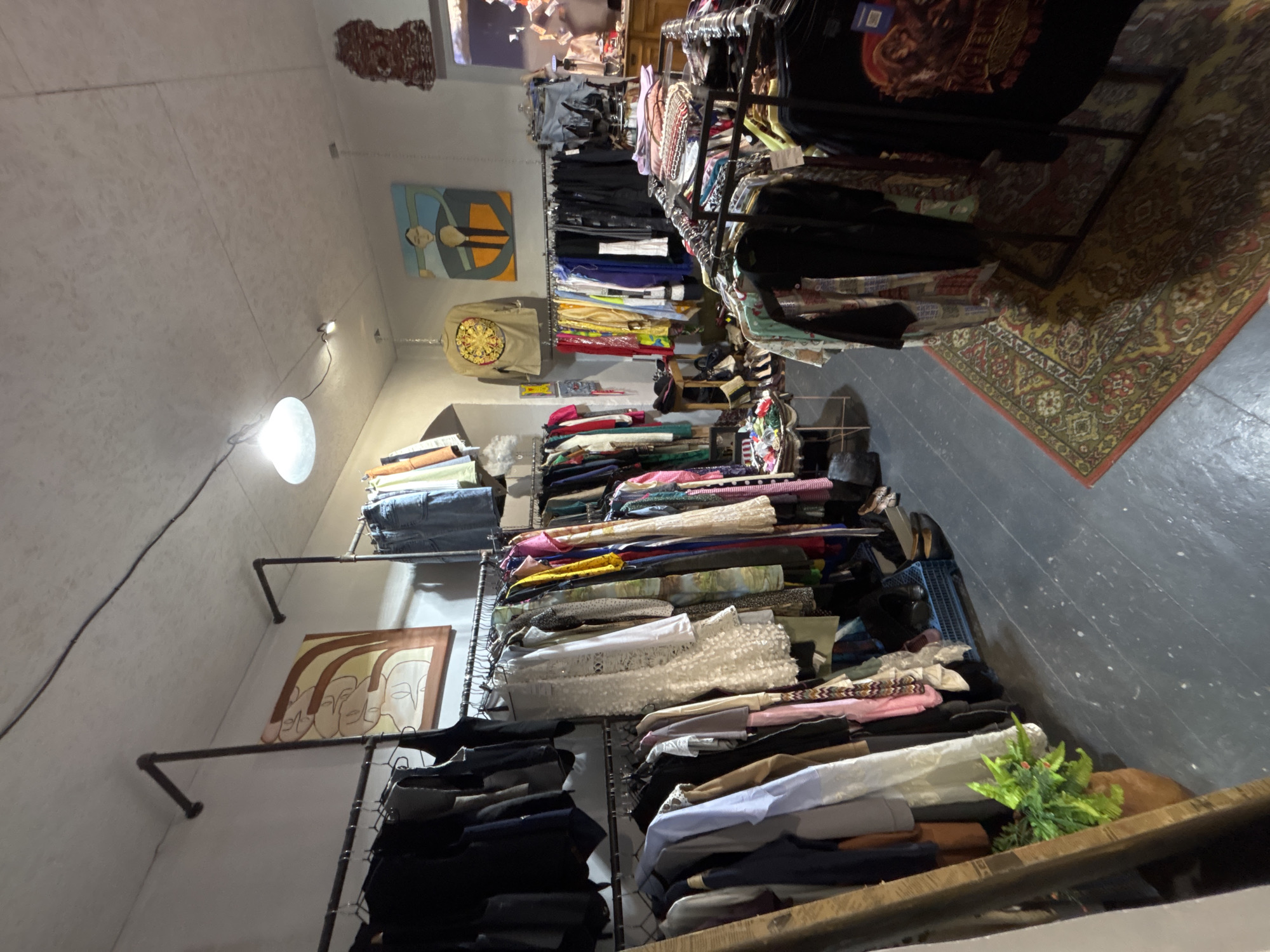 This image depicts the interior of a vintage clothing store, likely located in the city of Yerevan, Armenia. The scene is filled with racks of clothing, ranging from casual wear to more formal attire, all displayed on metal clothing racks. The store has a rustic and eclectic ambiance, with a concrete ceiling and dark wooden floors.
On the walls, there are several pieces of art, including an abstract painting with earthy tones and another colorful painting of a person. The lighting in the store is a mix of overhead lights, with one large, dome-shaped pendant light and smaller spotlights, creating a warm and inviting atmosphere.
In the foreground, there are several clothing racks displaying a variety of garments. The clothing appears to be mostly secondhand, with a mix of styles and colors. Some of the items visible include shirts, dresses, jackets, and jeans. A patterned rug with red and yellow accents is laid out on the floor in front of some of the clothing racks.
In the background, there is a wooden counter with various items displayed on it, suggesting a checkout area or a display for accessories. A small potted plant adds a touch of greenery to the space. The overall impression is one of a well-stocked and charming vintage boutique.