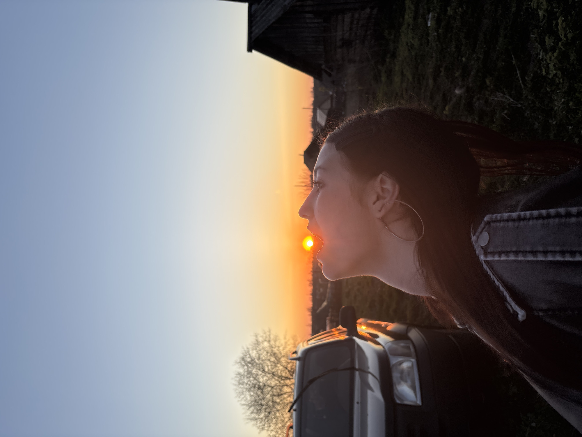 A woman with dark hair and wearing a large hoop earring and a dark jacket is photographed in profile. She is positioned so that the setting sun appears to be entering her mouth. The background includes a dark silhouette of a building, possibly a barn, and a tree without leaves. A vehicle is visible beneath the woman's chin, with headlights reflecting the sunset. The sky transitions from blue to orange near the horizon. The location might be near the city of Фоки, Russia, given the query context. The scene captures a playful interaction with the sunset, using the perspective to create a visually interesting effect.