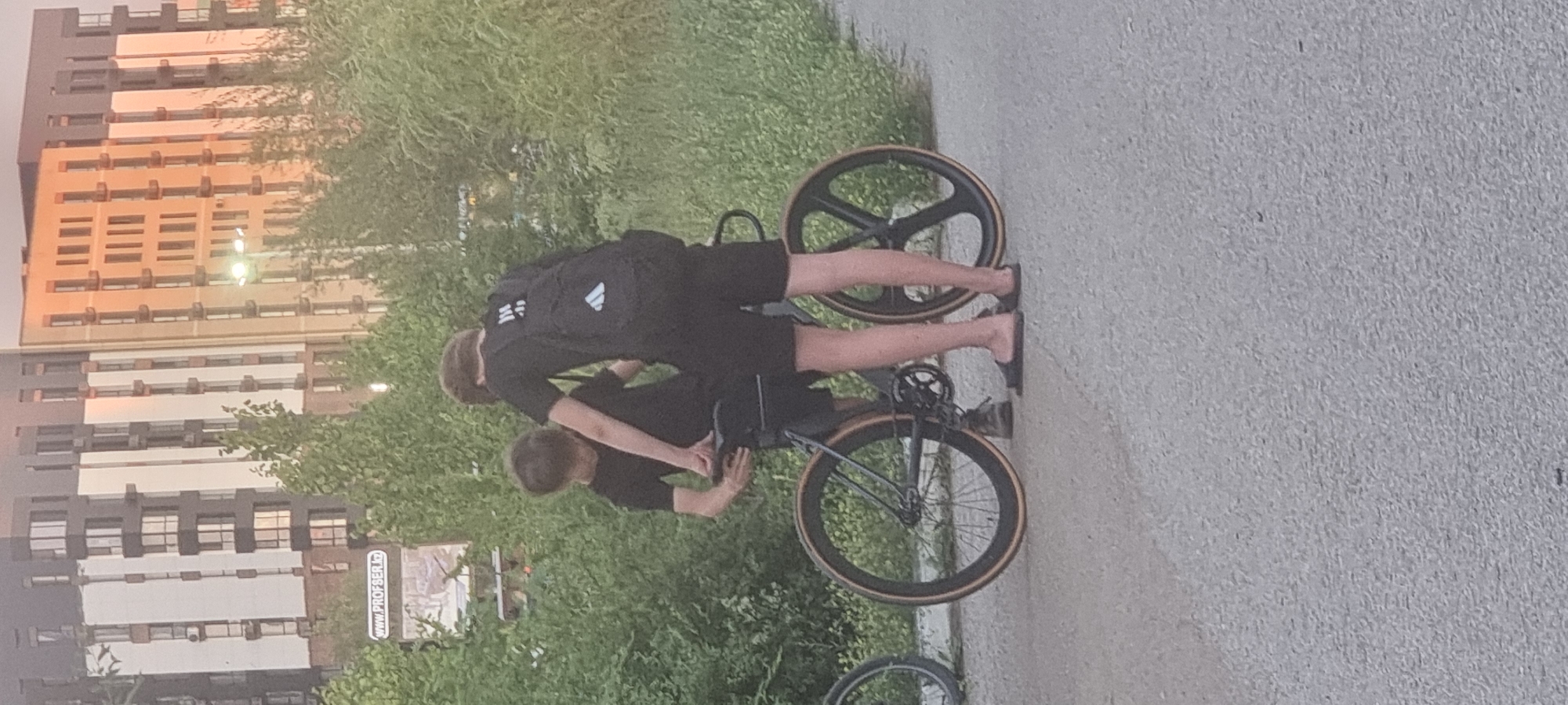 In Aktobe, Kazakhstan, two individuals dressed in black stand near a bicycle on a paved path. The person on the left is wearing a black t-shirt and shorts and is positioned by the bicycle's seat. The person on the right wears a black t-shirt with a backpack and black shorts. The bicycle has a black frame, one solid black wheel with a tan rim, and another wheel with a similar tan rim and a multi-spoke black design. In the background, lush green trees are visible, behind which stand two modern, multi-story buildings with numerous windows. One building has a light-colored facade with dark vertical accents, while the adjacent building is a darker beige or tan. A street lamp is positioned between the buildings. A sign with "www.PROFSER.kz" is affixed to a pole amidst the trees. The lighting suggests it is either early morning or late afternoon, with a warm glow illuminating the scene.