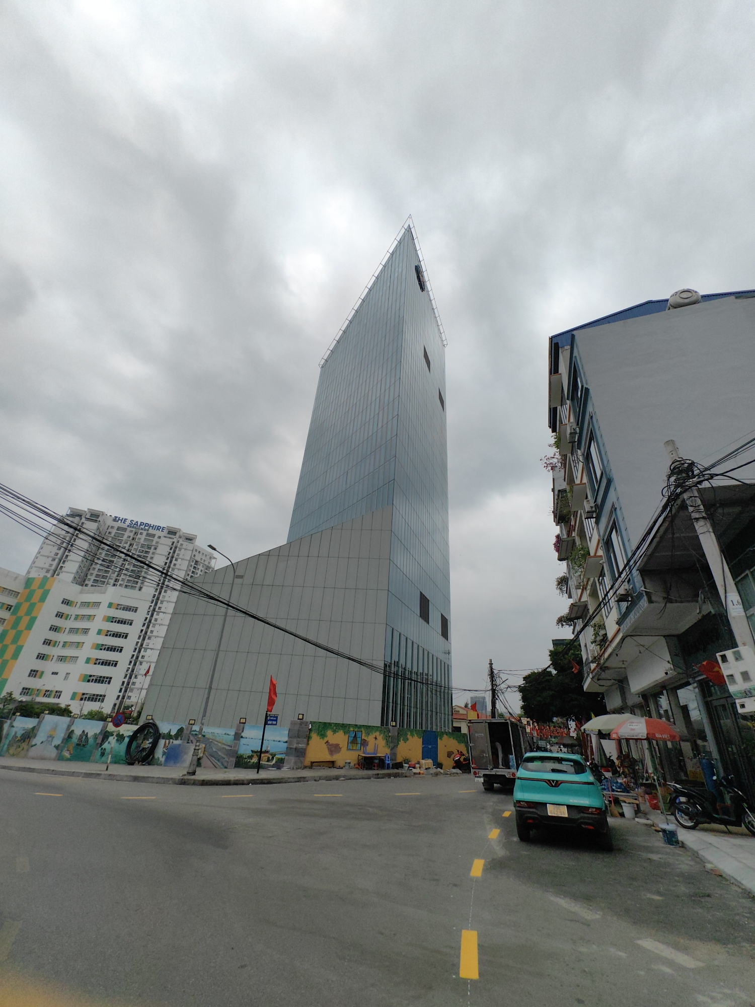 The image shows a street scene in Hong Gai Ward, Vietnam, under a cloudy sky. A tall, modern building with a glass facade dominates the view. To the left is another building with "THE SAPPHIRE" sign. A low concrete wall with murals runs along the left side of the street. The street is asphalt and has yellow traffic markings. A blue car is parked along the side of the road with other motorbikes near a street vendor with umbrellas. Power lines are visible overhead. A red flag is posted near the wall. Several buildings are visible behind the central tower.