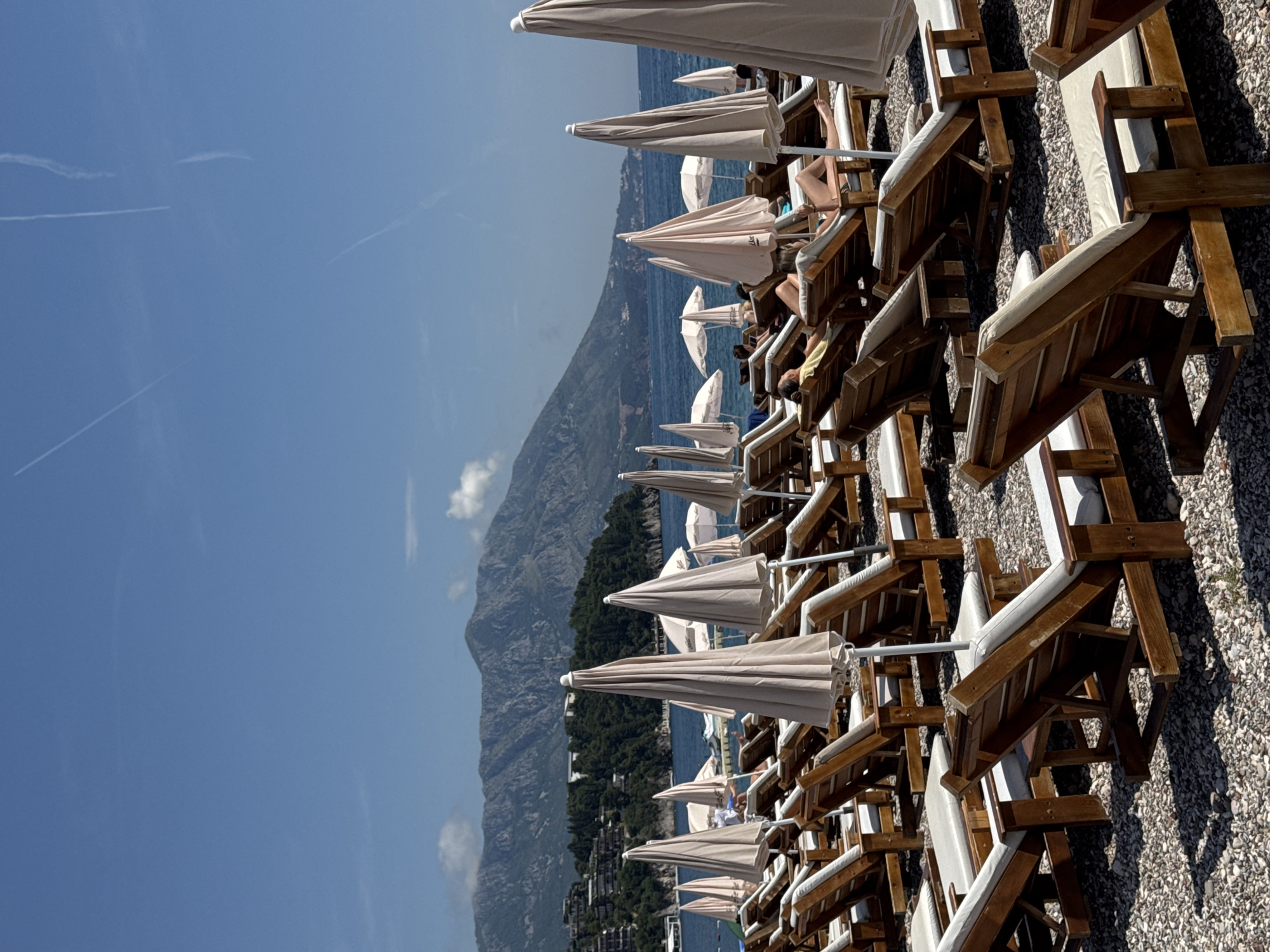 This image depicts a beach scene in Budva, Montenegro. The foreground features a row of wooden beach chairs with white cushions and closed beige umbrellas, arranged closely together on a pebble beach. Some chairs are occupied by people, presumably relaxing in the sun. In the background, a steep, forested mountain rises from the coast, forming a backdrop to the beach. The Adriatic Sea stretches out in front of the mountain, and the clear blue sky above contains thin contrails from aircraft. The overall impression is one of a popular and scenic beach resort.
