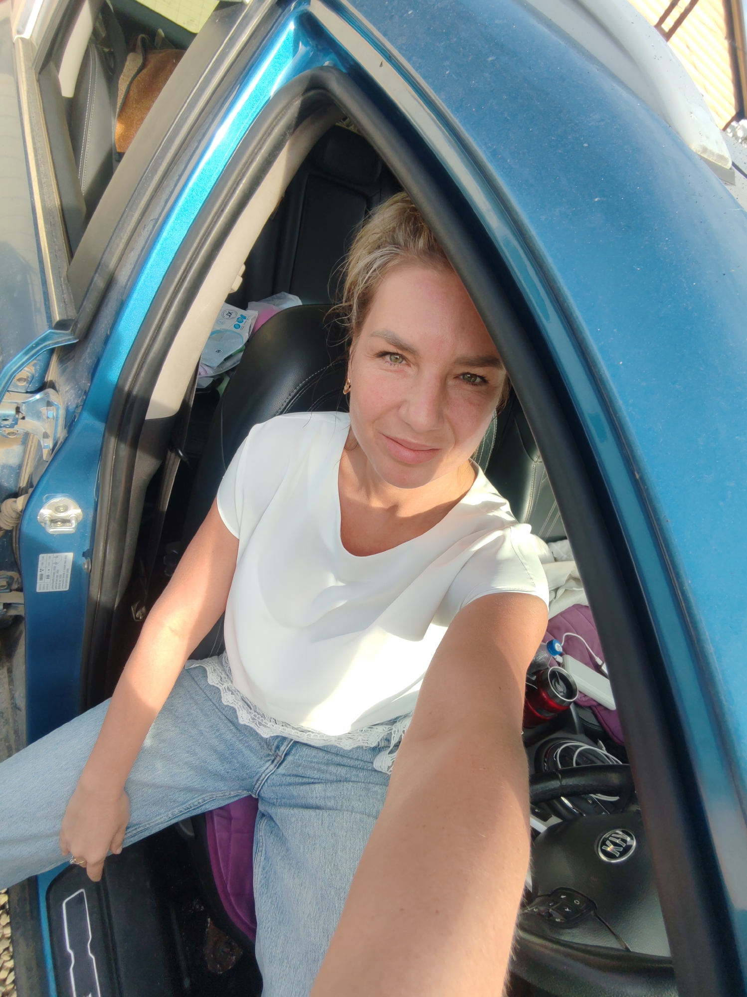 A woman with blonde hair pulled back is sitting in the driver's seat of a blue car. She is wearing a white t-shirt and light blue jeans. She is smiling at the camera. The car is parked outside, with sunlight illuminating the scene. The interior of the car is visible, including the steering wheel and dashboard. There are various items in the car, including a purple bag and a water bottle. The image appears to have been taken in Paratunka, Russia, as indicated by the context.