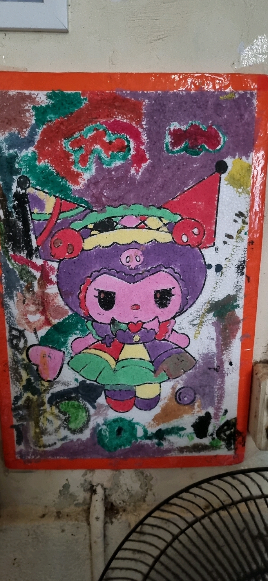 A framed artwork, possibly a drawing or painting, is affixed to a textured, off-white wall. The artwork features a stylized character with a large purple head, pink face, and black eyes. The character is wearing a colorful, patchwork-style outfit with a green skirt. It has a festive or circus-like headdress with red cones and skull-like decorations. The background of the artwork is a chaotic blend of colors, including red, purple, and green, with a textured, speckled appearance. The artwork is bordered by a bright orange frame. The wall itself appears worn, with visible texture and some discoloration, possibly indicating age or environmental damage. To the right of the artwork, a portion of a white electrical conduit is visible, along with what appears to be a part of a black metal fan grille in the lower right corner of the image. The scene suggests an indoor setting, potentially a home or a public space with artistic elements. Given the context of Denpasar, Indonesia, this could be a personal display in a residence or a piece of local art in a community area.