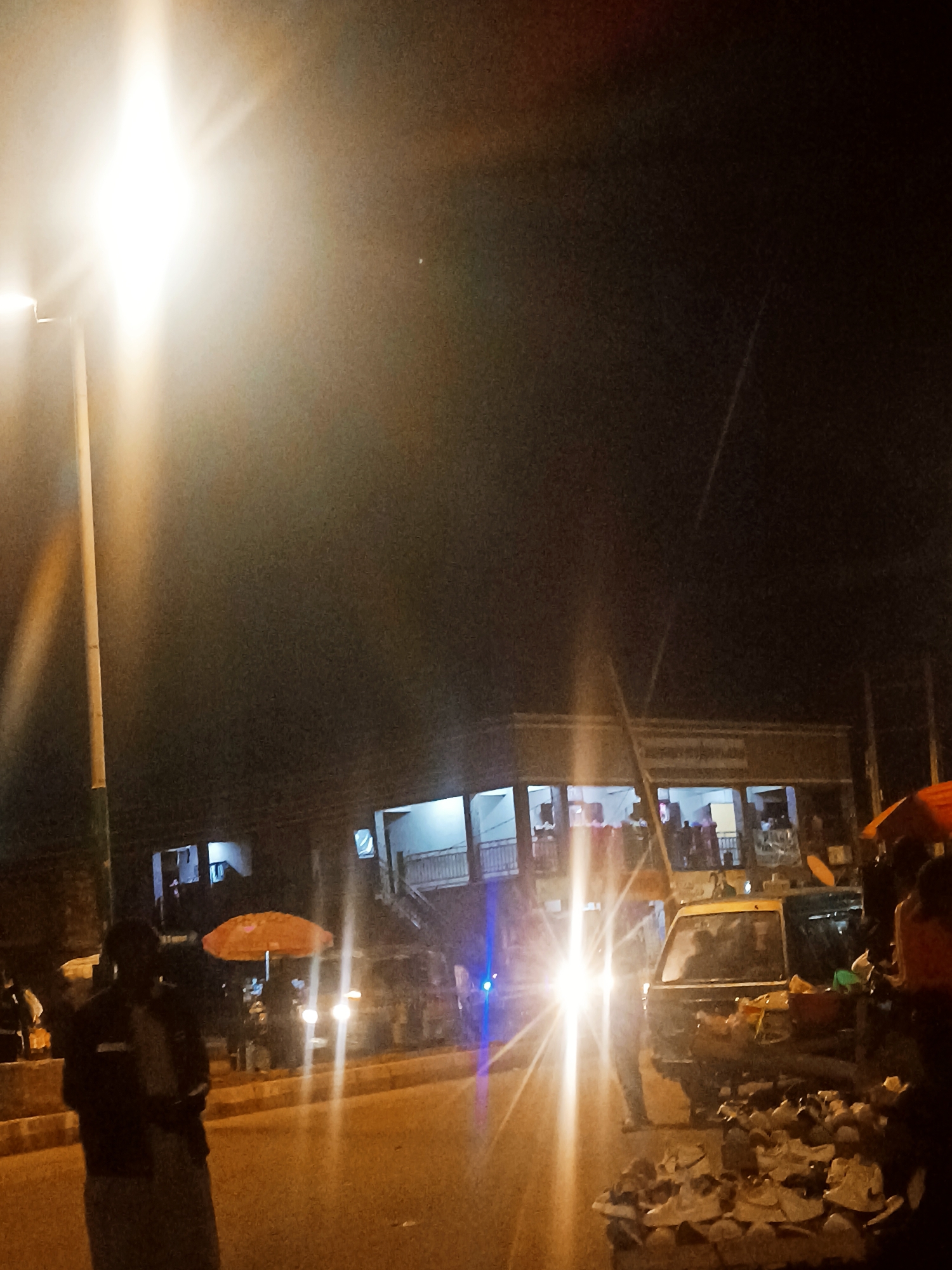 The image depicts a street scene in Bukuru, Nigeria, at night. A streetlight on the left illuminates the foreground. In the middle ground, a two-story building with open railings and visible people on the upper level is present. Car headlights are a central focal point, creating bright streaks. To the right of the headlights is a parked vehicle resembling a taxi, surrounded by people. A stall with merchandise, possibly footwear, is in the foreground. A man stands to the left, wearing a dark jacket. An umbrella with orange fabric is visible.