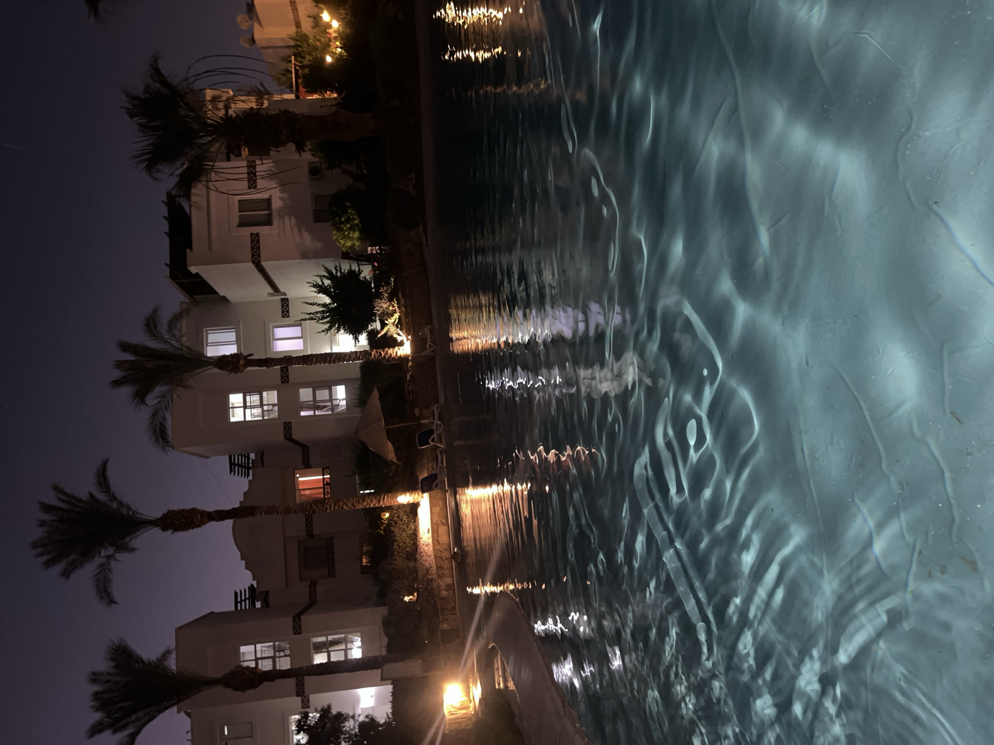 This nighttime image depicts a resort swimming pool area in Sharm El Sheikh, Egypt. The foreground is dominated by the rippling surface of the pool, reflecting the ambient light from nearby sources. In the background, several white buildings with illuminated windows are visible, interspersed with tall palm trees. Some of these buildings appear to be hotels or residential units, typical of a resort setting. Lounge chairs and a parasol are present by the poolside, suggesting a recreational area. The sky is dark, indicating it is late evening or night, and a soft, warm glow emanates from some of the artificial lights, creating reflections on the water. The overall atmosphere is tranquil and inviting.