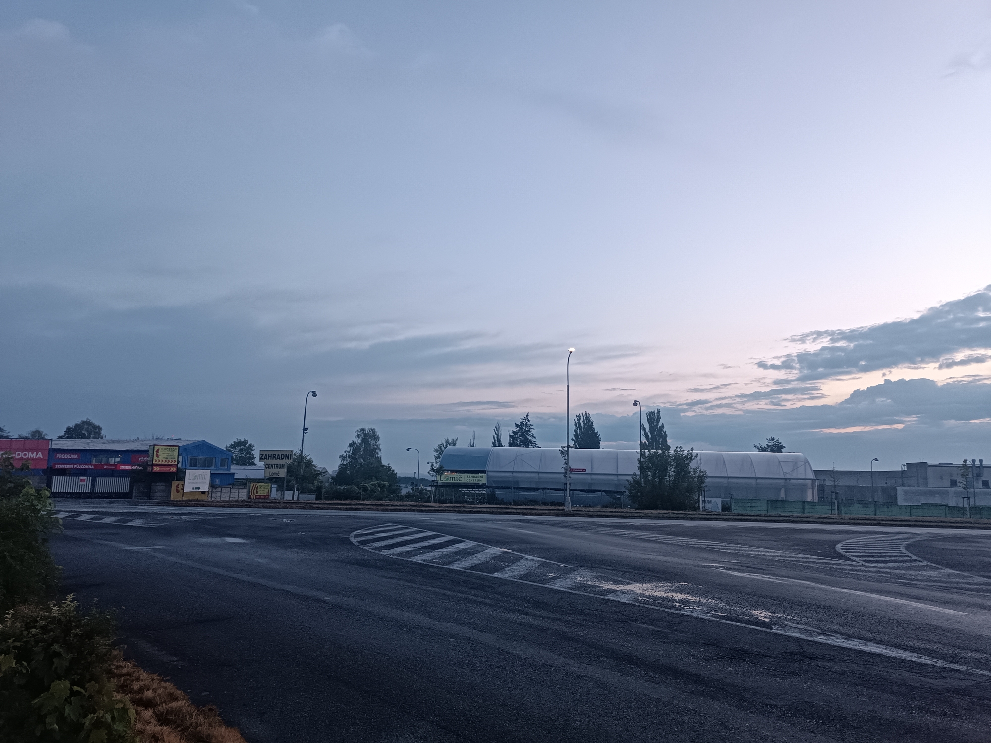 The image captures an exterior view of a retail and industrial area in Přelouč, Czechia, during what appears to be early morning or late evening, given the subdued lighting and overcast sky. The scene is dominated by a wet asphalt road in the foreground, suggesting recent rain. Road markings, including a speed bump and a circular lane divider, are visible.

To the left, a row of commercial buildings lines the street. One building features large banners, one of which reads "DOMA" in red and white, and another displays "PRODEJNA" and "STAVEBNÍ PŮJČOVNA". Adjacent to these, a blue building with "ZAHRAHNÍ CENTRUM LOMC" signage is visible. Further down the street, industrial structures with white coverings, possibly greenhouses or temporary structures, are present, accompanied by trees and a street lamp. A chain-link fence runs along the front of some of these structures.

The sky is a pale blue with scattered clouds, some appearing darker and more textured, particularly on the right side. No people are present in the frame. The overall impression is one of an urban commercial zone at a quiet time of day.