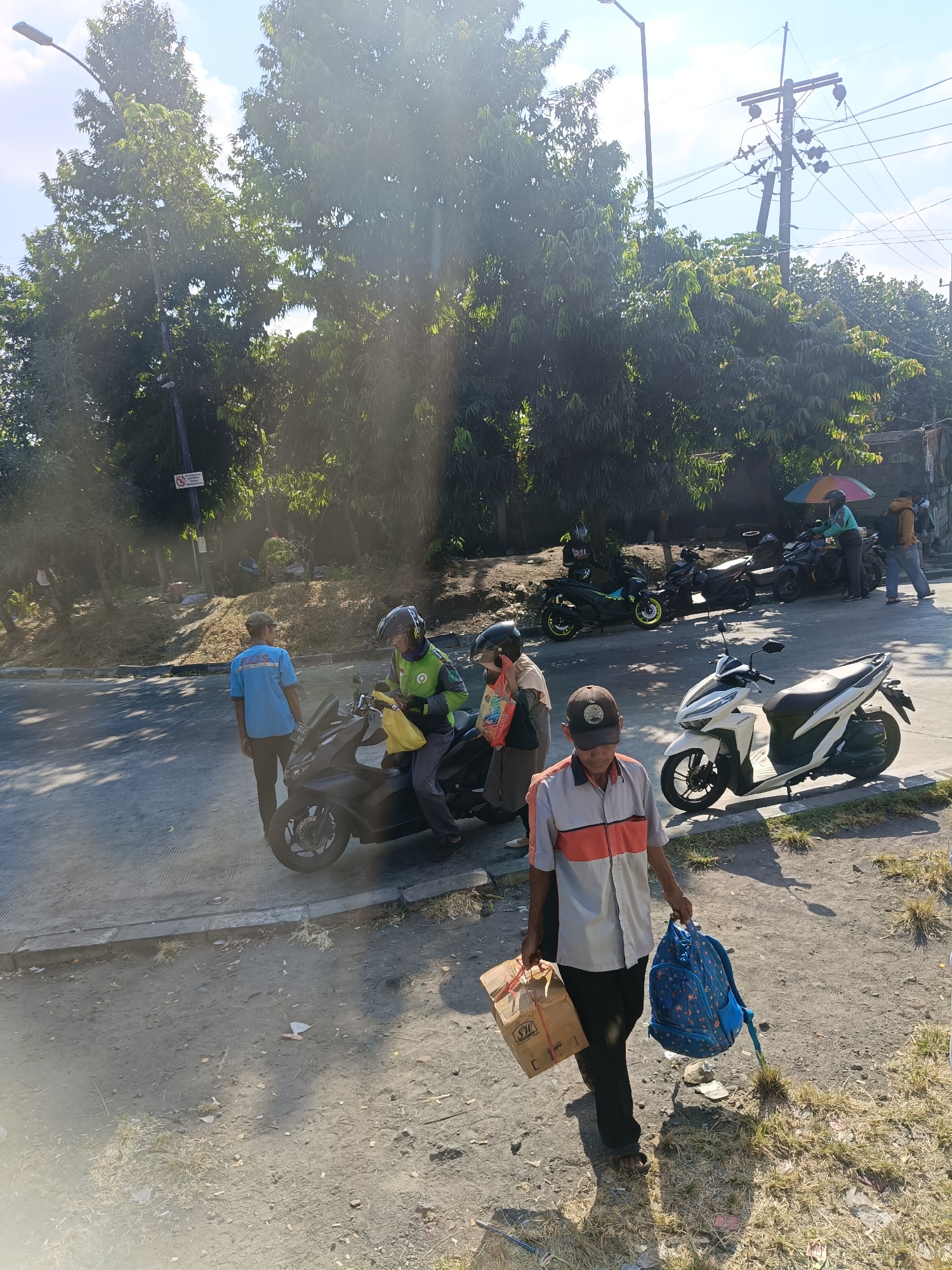 In Kedungrejo, Indonesia, a man in a striped short-sleeved shirt and dark pants walks on a dusty, sunlit road. He carries a brown cardboard box tied with string in his left hand and a blue backpack with a floral pattern in his right hand. To his left, a white motorcycle is parked, and further back, a dark scooter is ridden by a person in a green jacket. Several other motorcycles are parked in the background, some under a colorful umbrella. Trees line the edge of the road, and power lines are visible overhead. Another man in a blue shirt with writing on the back stands near the scooter. The scene suggests a busy roadside environment, possibly a transportation hub or a commercial area.