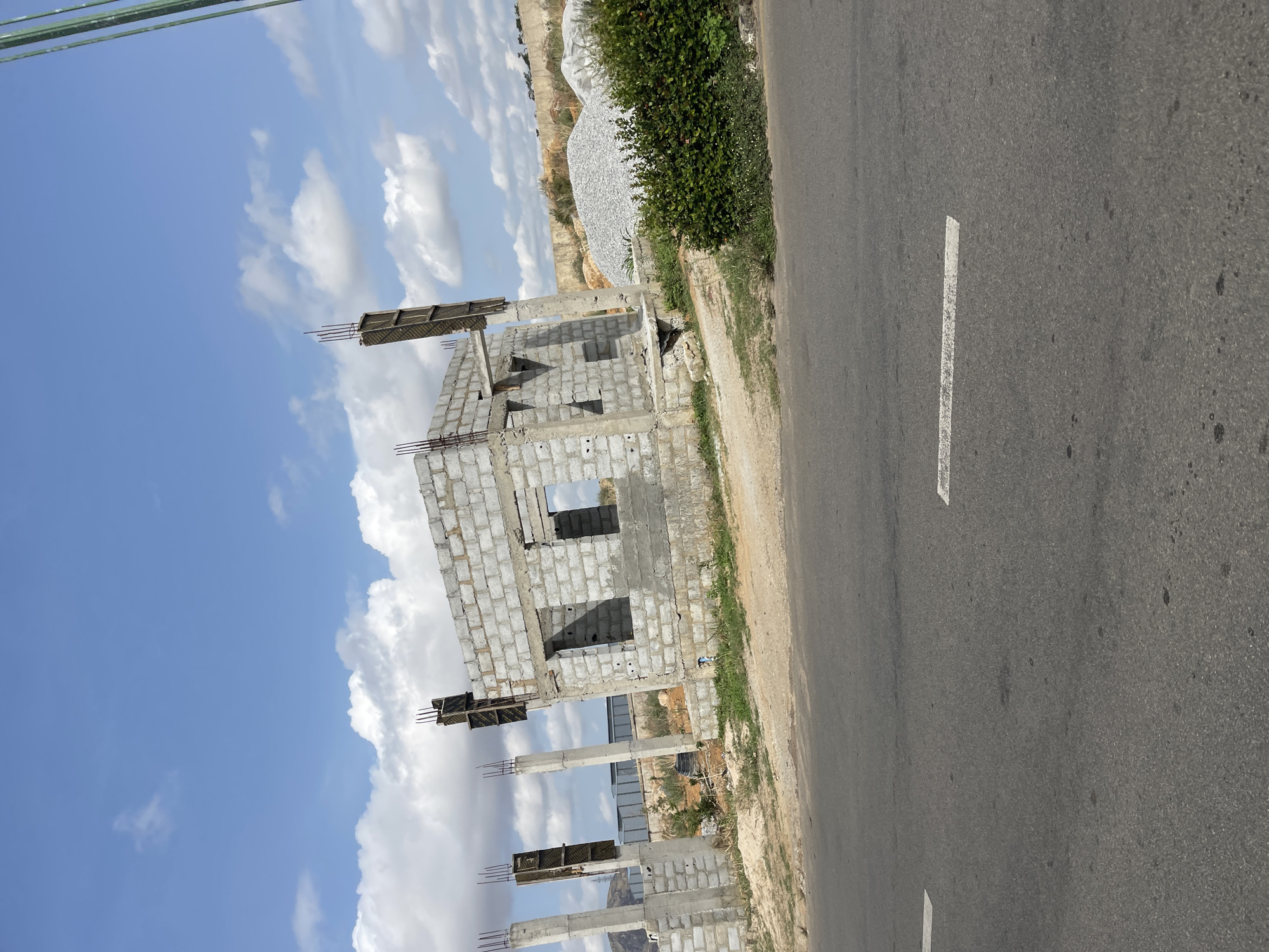 The image shows a building under construction in Gosa, Nigeria. The structure is made of concrete blocks and appears to be a multi-story building. The construction is incomplete, with exposed rebar and partially built walls. The building sits close to a paved road with a white dividing line. Adjacent to the road is a strip of land with grass and some shrubbery. In the background, there's a pile of gravel or stones near a mound of dirt, possibly indicating a construction materials storage area. The sky is visible with scattered clouds.