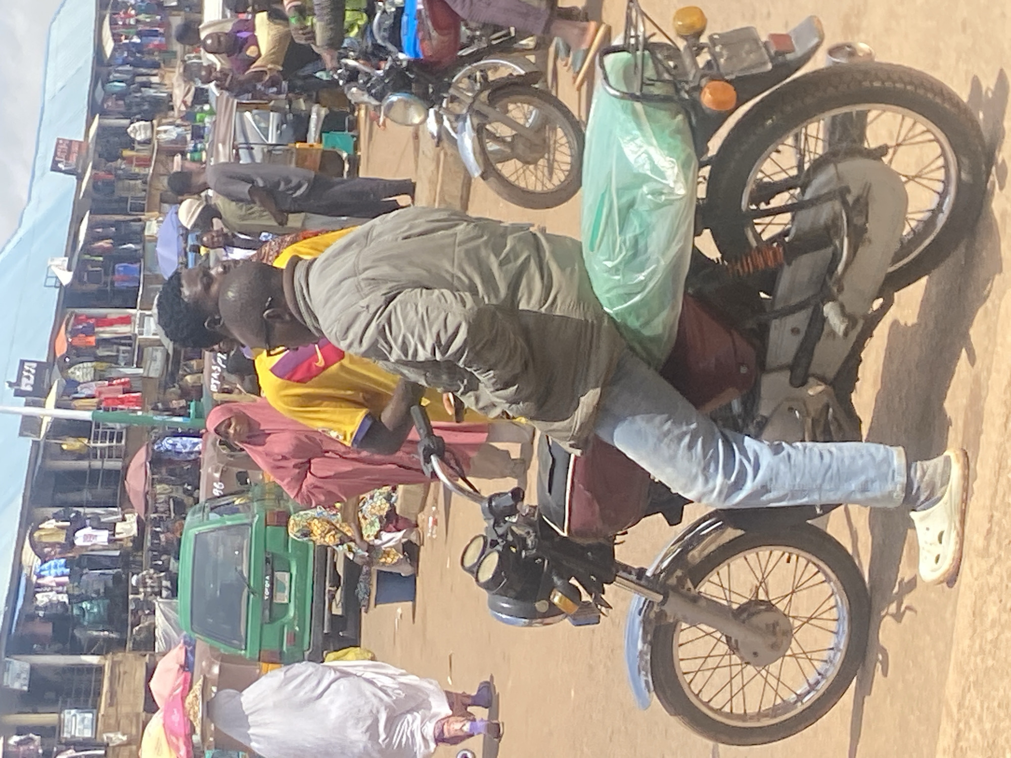 The image depicts a vibrant marketplace scene in Bukuru, Nigeria. The primary focus is on a man on a motorcycle, who appears to be in motion or preparing to move. He is wearing a grey jacket and light blue jeans, with white crocs on his feet. The motorcycle is a dark color with a silver fender and spoke wheels. A large, crinkled, light green plastic bag is strapped to the back of the motorcycle.
In the background, a bustling market is visible with stalls selling various goods, including clothing, bags, and what appear to be electronic items. Several other people are present, some walking, others gathered around stalls. A green, three-wheeled vehicle, possibly a commercial tricycle or "Keke Napep," is parked nearby. A woman in a pink headscarf and yellow shirt is visible in the mid-ground. The overall impression is of a busy, sunny day, typical of an outdoor market in West Africa. The lighting suggests it is daytime, likely mid-morning or afternoon. The activity appears to be commerce and daily life in a busy urban environment. There is some text visible on signs and the green vehicle, but it is too blurry to read clearly. The atmosphere seems lively and active.