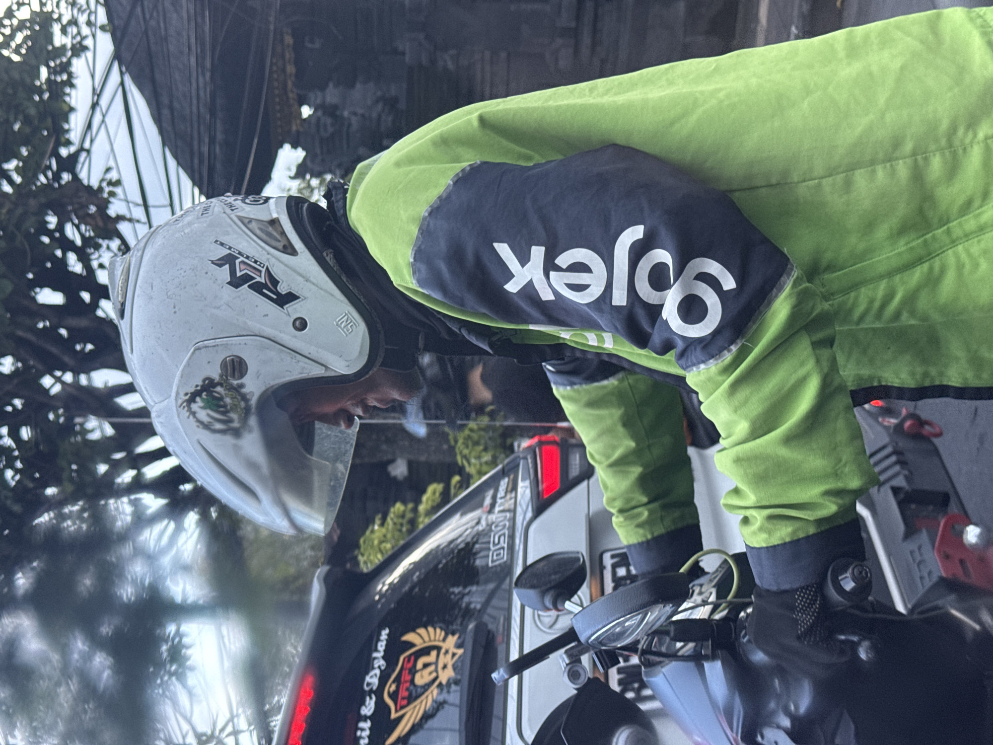 The image, captured in North Kuta, Indonesia, depicts a person on a motorcycle, likely a ride-hailing driver, paused on a street during the daytime. The individual is viewed from a side-rear perspective, wearing a distinctive bright lime green and dark blue jacket, with the "Gojek" logo prominently displayed in white on the dark blue sleeve (appearing reversed due to the angle). They are also wearing a white full-face helmet featuring "RY" branding, the word "HELMET," and an "SNI" (Indonesian National Standard) certification sticker. A wisp of smoke near their face indicates they are smoking while waiting.
The rider's hands rest on the motorcycle's handlebars, which are partially visible. In the background, a dark-colored car is present, featuring a rear window sticker with a shield-like logo that includes the text "TRFC" and the names "ANIL & DYLAN," along with "DSWR" on another part. Another vehicle is visible further back. The lighting suggests it is daytime, possibly overcast or with diffused light. Blurred trees and some distant architectural elements frame the background. The scene captures a moment of rest or waiting amidst daily activities in an urban or semi-urban setting.