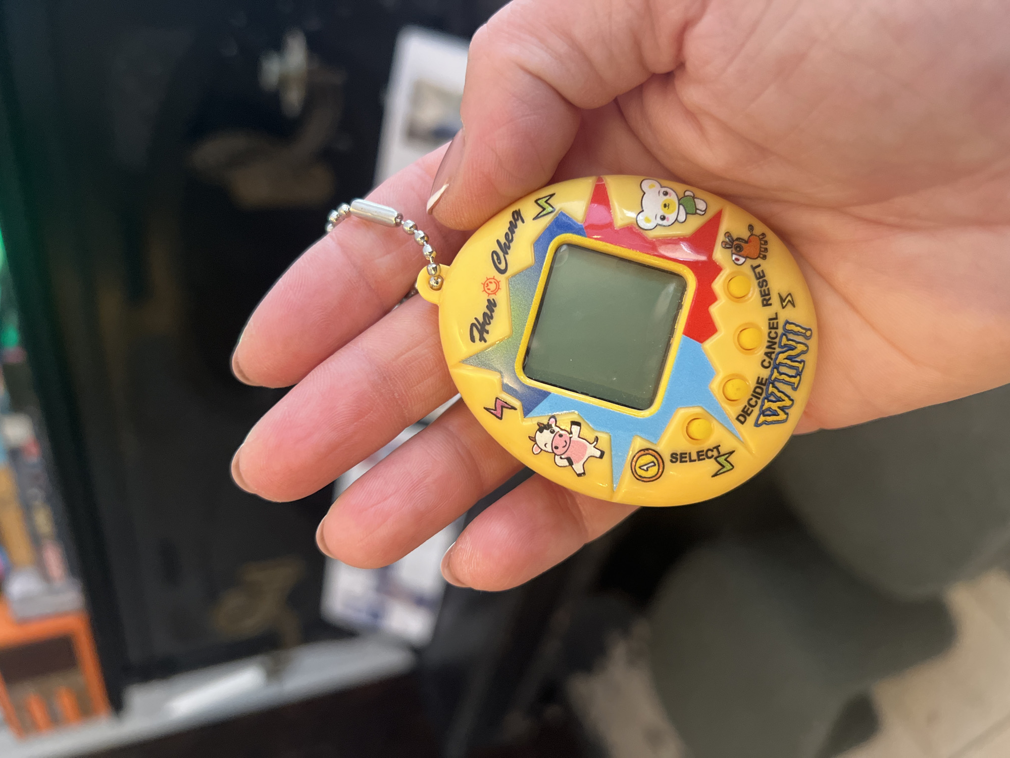 A person's hand holds a yellow, egg-shaped electronic pet toy, a Tamagotchi. The toy is decorated with cartoon characters, including a cow and a bear, and text that reads "Han Cheng," "DECIDE CANCEL RESET," and "WIN!". The toy has a screen and several buttons. The background is blurred but appears to be indoors, possibly a room with furniture and shelves. The location of this image is unknown and cannot be determined from the visual information provided.