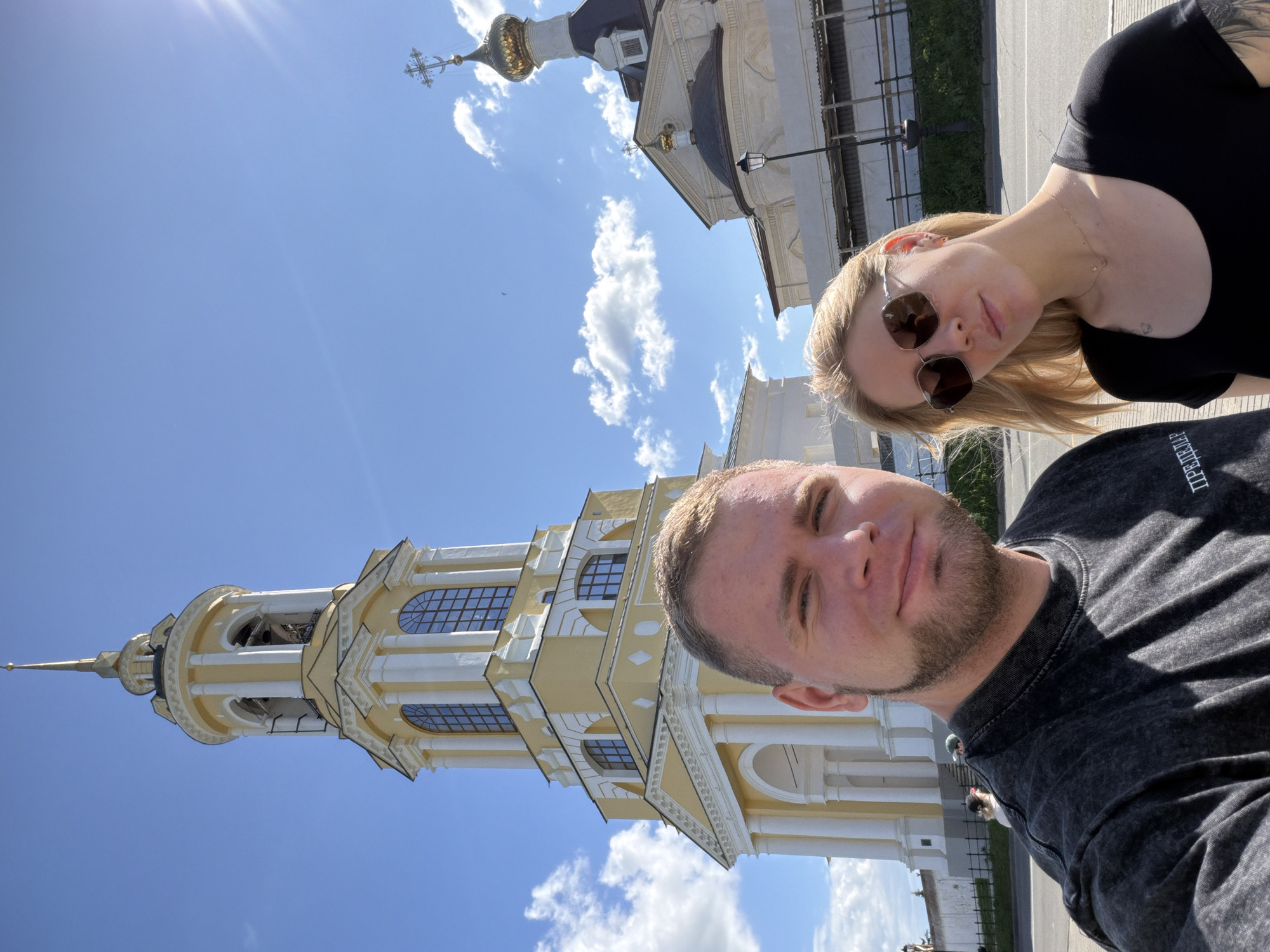 A selfie is taken by a man and a woman in front of a yellow bell tower and a domed building with golden crosses. The man, with a beard and short hair, is on the left, wearing a dark grey t-shirt. The woman, with blonde hair and sunglasses, is on the right, wearing a black t-shirt. The bell tower, a prominent architectural feature, is tall and slender with arched windows and decorative white elements. Behind it, a building with a dark roof and a golden dome is visible. The sky is blue with scattered white clouds. The scene is set in Suzdal, Russia, a city known for its historical churches and monasteries.