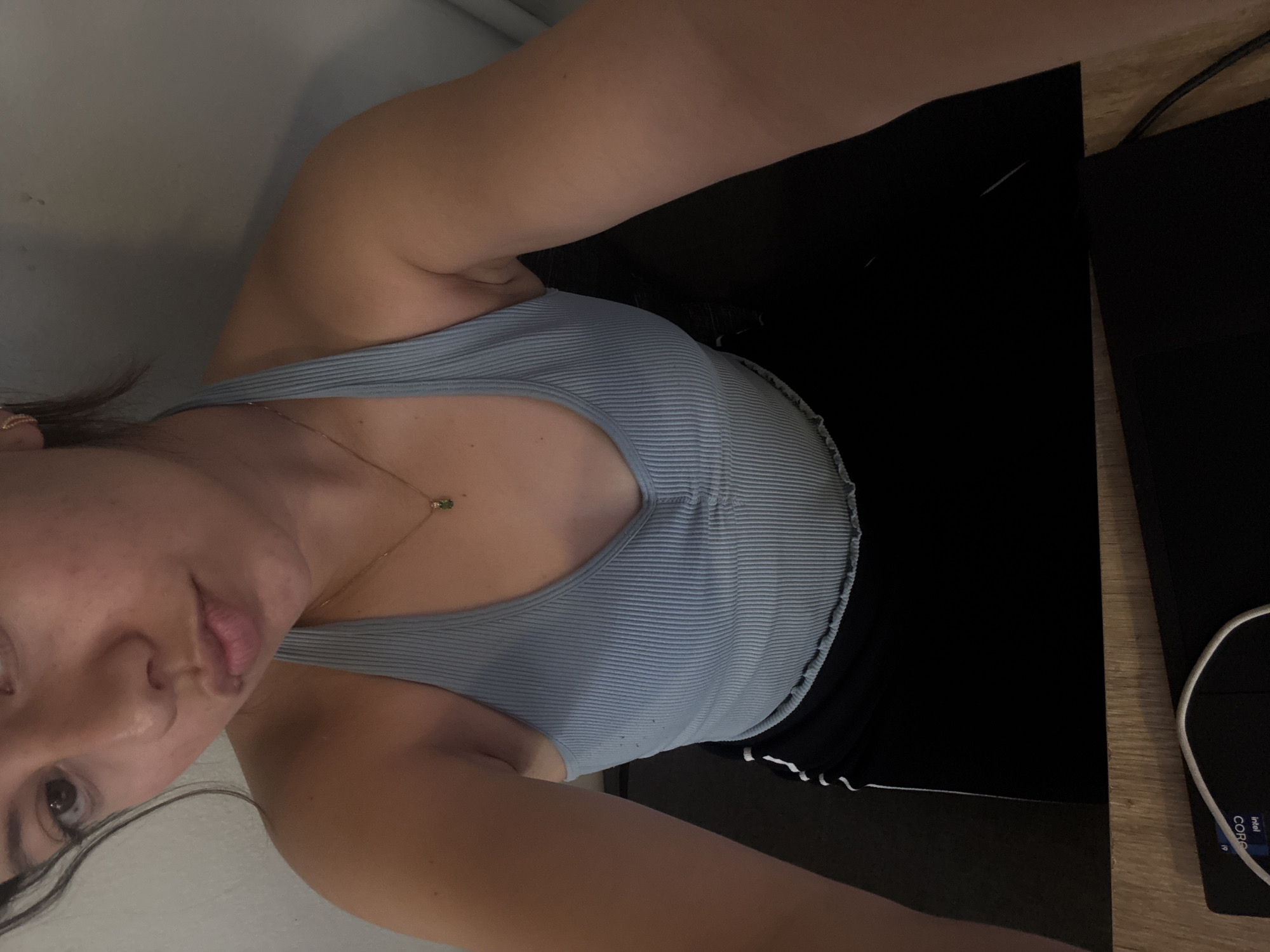 A selfie of a woman indoors. She is wearing a ribbed light blue tank top and black bottoms with white trim. She is wearing a gold necklace with a green pendant. A black laptop and white charging cable are on a wooden surface to her right. The walls are a light neutral color. No details indicate the location is San Andrés Cholula, Mexico.