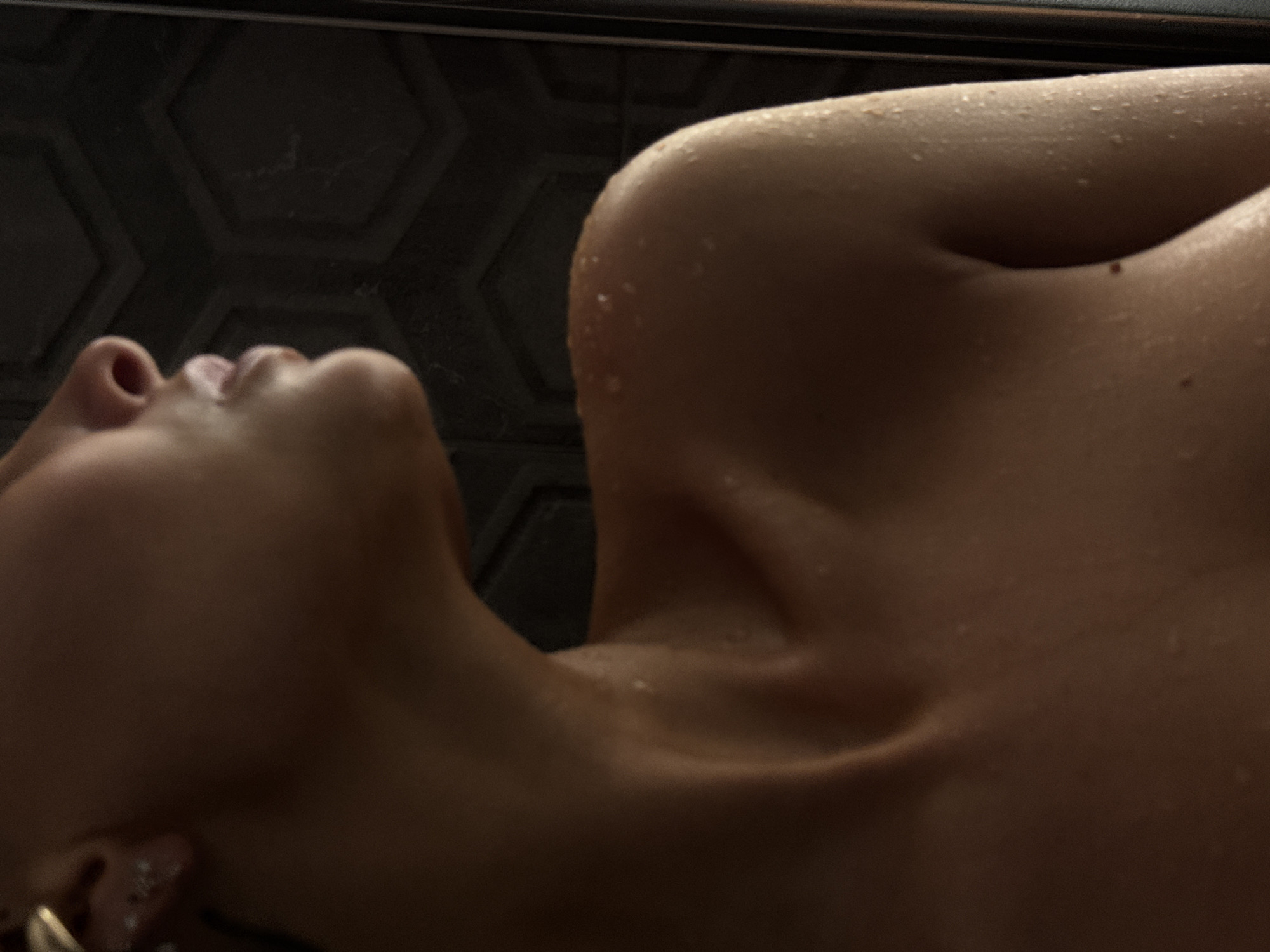 The image is a close-up, low-angle shot of a person's upper body, focusing on their neck, shoulder, and partial torso. The subject is partially submerged in water, evidenced by water droplets on their skin and the wet appearance of their shoulder and upper chest. The lighting is dim, casting shadows and highlighting the contours of the body. The background features a textured dark wall, possibly in a bathroom setting, with a hexagonal pattern. The person's face is not fully visible, with only a profile of their nose and mouth visible in the upper left of the frame. There is an earring visible on the left earlobe. There are no discernible landmarks or elements within the image that would indicate a specific location in Chaikovsky, Russia.