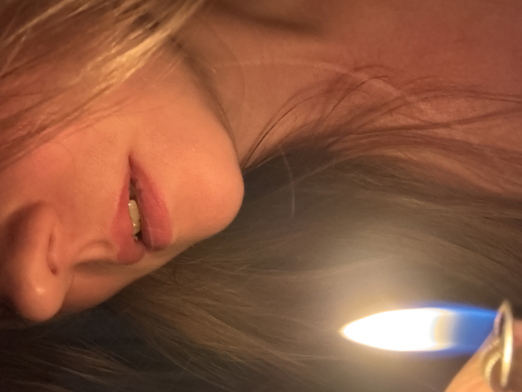The image shows a close-up of a woman's face illuminated by a lighter. The woman has fair skin and blonde hair. Her lips are slightly parted, revealing a tooth. The lighter flame is bright and blue-ish white and casts light on her face and hair. The background is dark and out of focus, suggesting a dimly lit environment. The location of the image is not identifiable from the provided details and there is no indication that it was taken in the city of Chaikovsky, Russia.