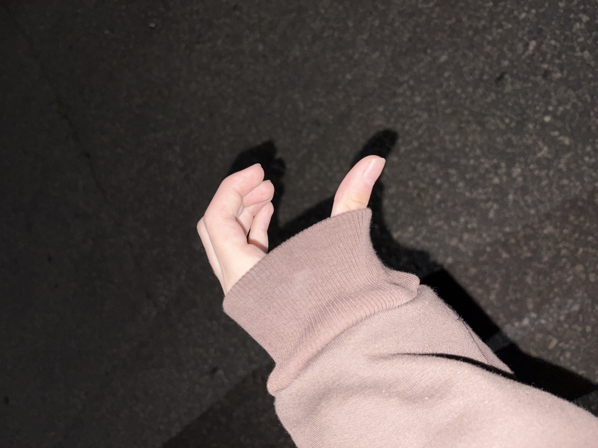 A close-up shot focuses on a person's left hand forming the shape of a heart. The hand is partially covered by the sleeve of a light pink sweatshirt. The background is dark and textured, appearing to be pavement. The scene is lit by an unseen light source casting a shadow of the hand. The location, Відзаўскі сельскі Савет, Belarus, is noted, but there are no visual cues in the image to confirm this specific location.