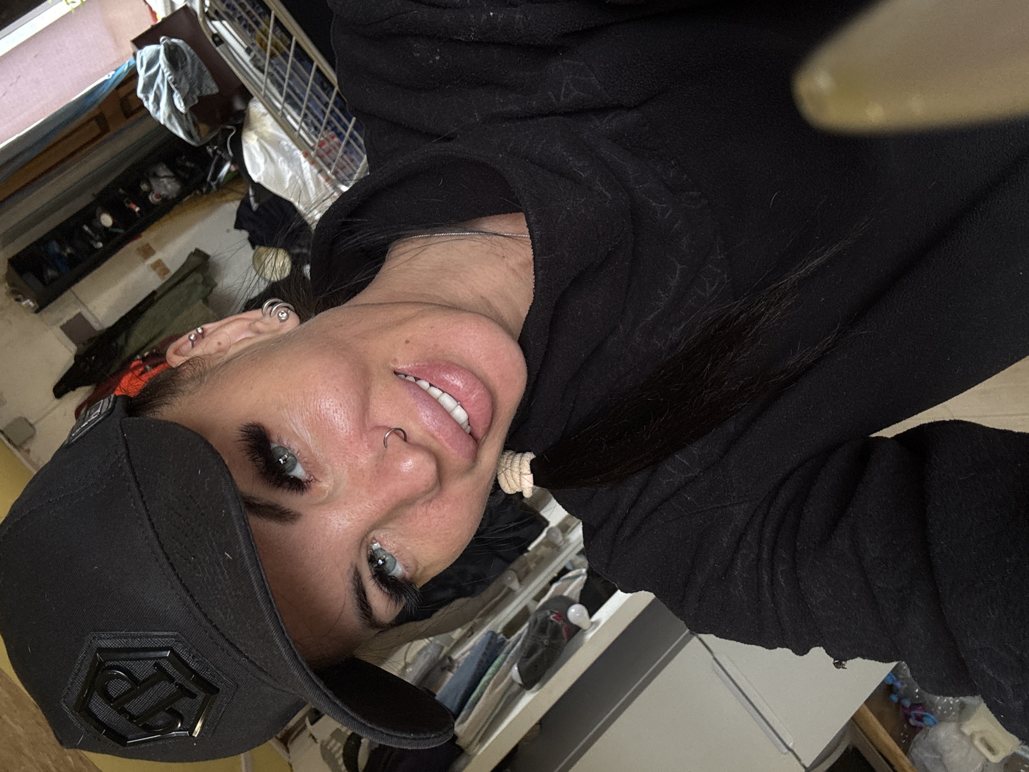 A woman, wearing a black baseball cap and a black hooded sweatshirt, is taking a selfie indoors. She has blue eyes, a nose ring, and visible eyelashes. Her hair is pulled back in a ponytail. The background appears to be a cluttered room with shelves filled with various items, including clothing and household goods. The lighting suggests it is daytime. There is no identifiable information about the location being Dulepovo, Russia.