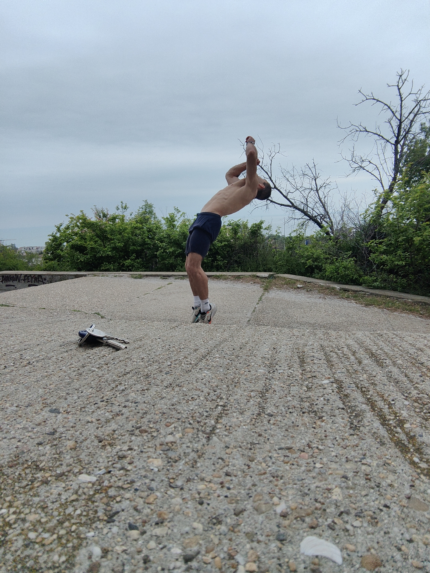 A person with a bare torso and dark shorts is performing a backbend on a gray concrete surface surrounded by green bushes and trees. The person is wearing athletic shoes with white socks. The sky is overcast. A set of keys lies on the concrete surface. A bare tree branch is visible in the background. The location is likely in Varna, Bulgaria, as indicated by the graffiti on the concrete edge.