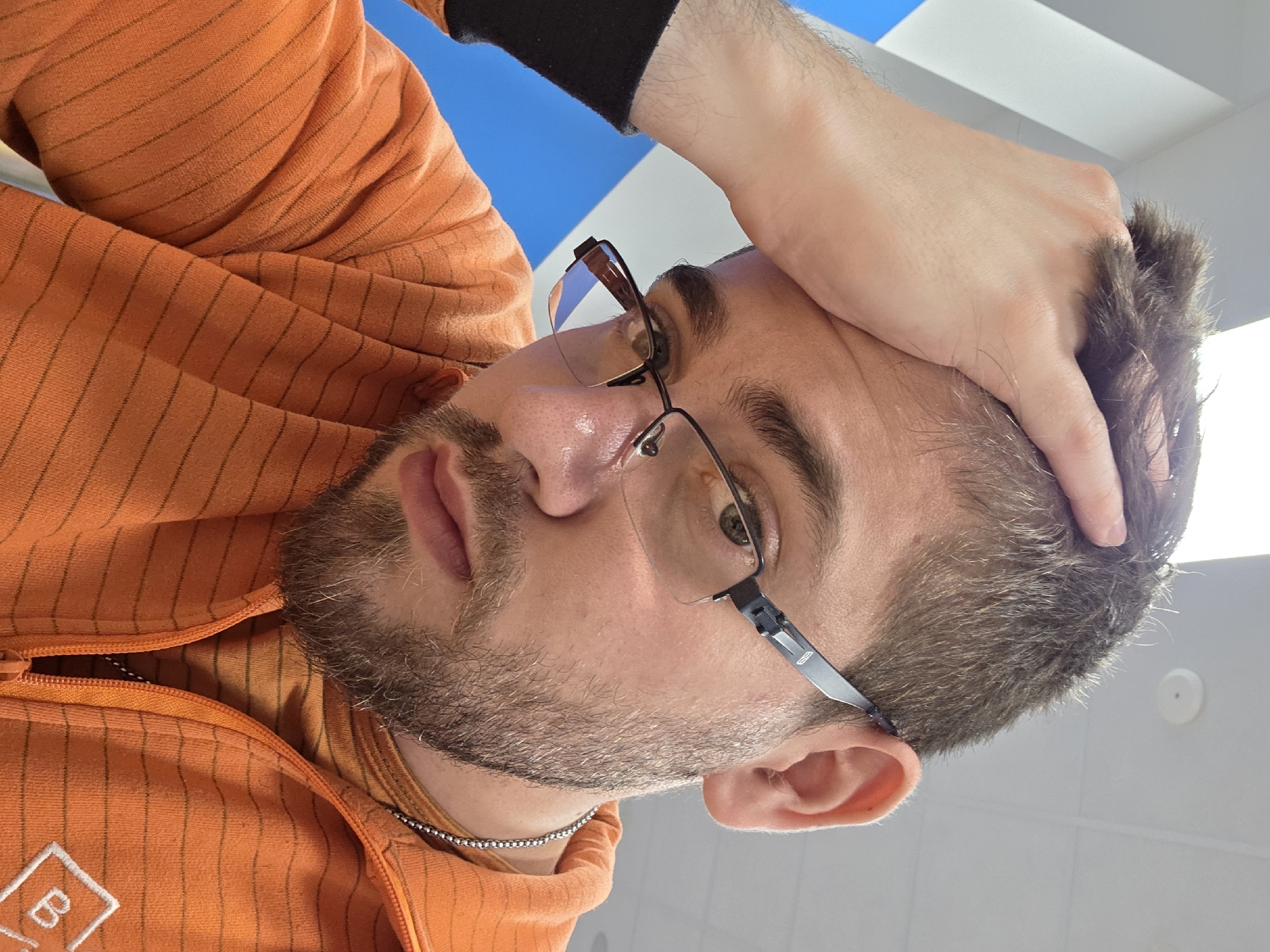 A man with a beard and glasses is looking at the camera with his hand on his head. He is wearing an orange striped jacket and a black sleeve can be seen at the top. He is also wearing a silver chain necklace. The background appears to be a blue sky and a white ceiling. The image was most likely taken indoors. It is impossible to verify the location to be Plovdiv, Bulgaria.