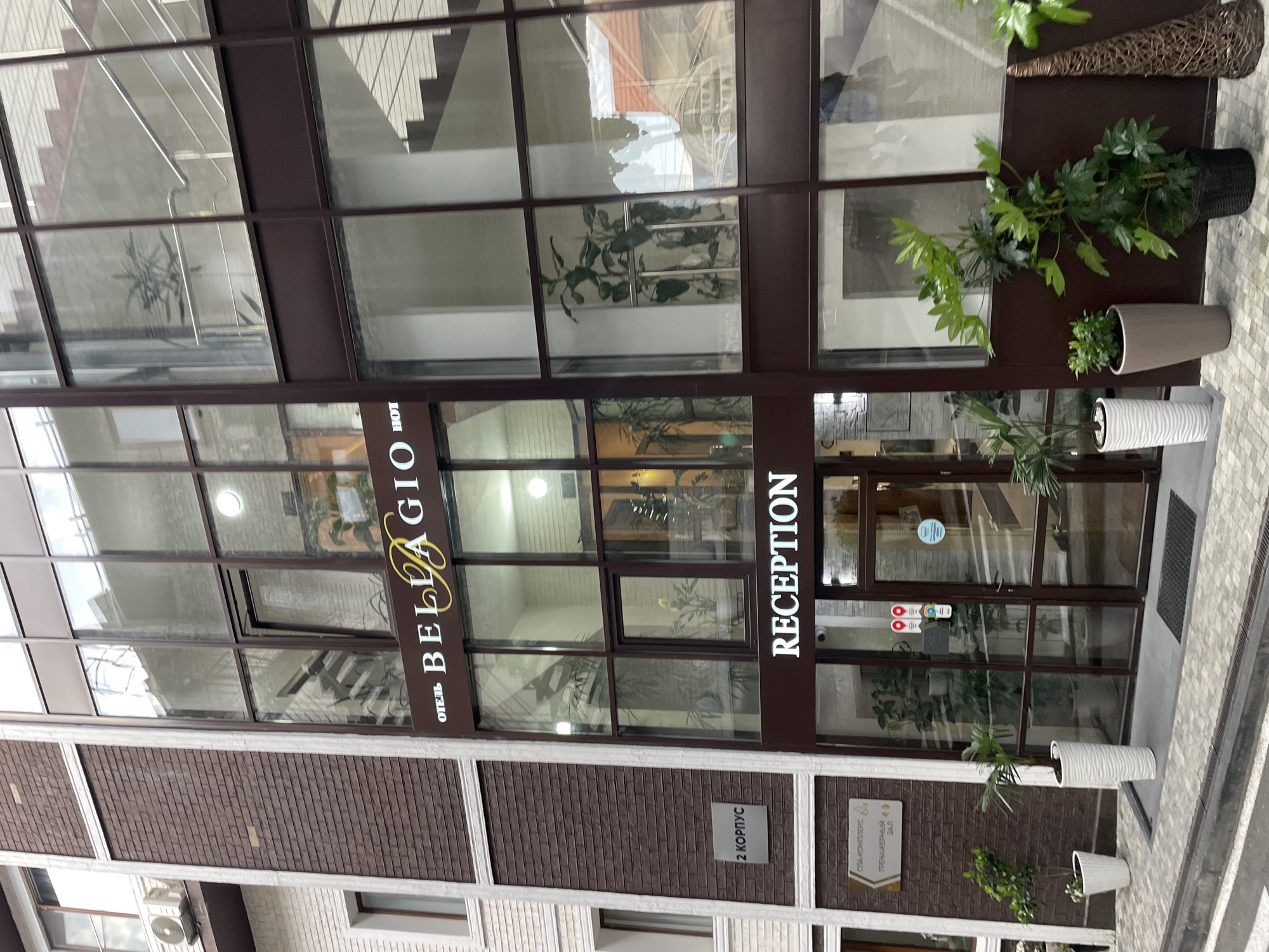 The image shows the exterior of Hotel Bellagio in Sochi, Russia. The building has a modern design with a large glass facade framed by dark brown metal. The word "RECEPTION" is prominently displayed in white lettering, adjacent to the entrance. Planters with greenery decorate the facade. The building is a combination of glass, metal, and textured brown brick.