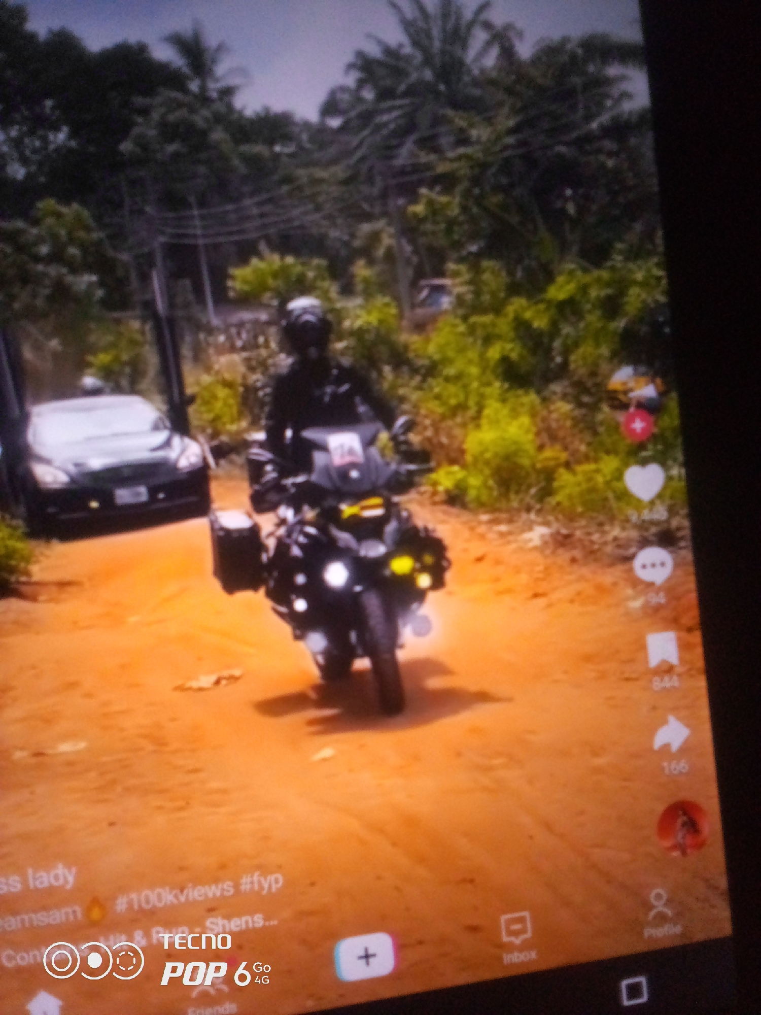 The image captures a TikTok video interface overlaying a scene of a person riding a motorcycle on a dirt road. The rider is wearing a helmet and dark clothing, and the motorcycle is equipped with storage boxes. To the left of the motorcycle, a black car with its doors open is parked on the side of the road. The environment appears to be a rural or semi-rural area with lush green trees and vegetation in the background. The road is unpaved and appears to be made of reddish-brown dirt. The TikTok interface elements visible include a heart icon with a count of "9,446", a speech bubble icon with "94", and a bookmark icon with "844". There are also icons for "Inbox" and "Profile" at the bottom of the screen. Text overlays indicate hashtags such as "#100kviews" and "#fyp". The overall scene suggests a travel or adventure-themed video. The location is identified as Howayi, Nigeria, though this context is not directly evident from the visual information alone.