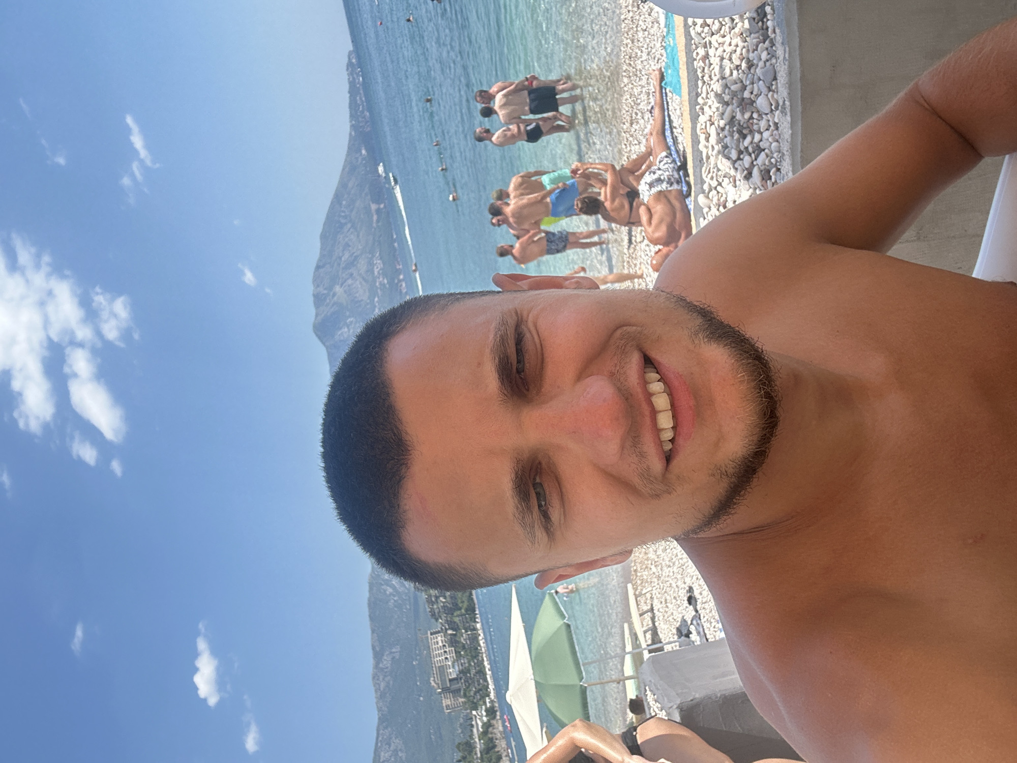 This is a selfie taken at a beach in Budva, Montenegro. The main subject is a young man with short dark hair and a beard, smiling at the camera. He is shirtless and appears tanned.
In the background, a crowded beach scene unfolds under a bright blue sky with scattered white clouds, suggesting it is a sunny day. Several people are visible, some wading in the clear blue water, others standing on the pebble beach. The water is calm, with a few small boats or jet skis in the distance. Buildings are nestled into the hillsides behind the beach.
The setting includes beach chairs and umbrellas, one of which is green and white. The shore is made up of small white and gray pebbles. The man's arm and chest are prominent in the foreground, also showing his tan. The overall mood is relaxed and vacation-like, typical of a summer day at the beach. No visible text is present in the image.