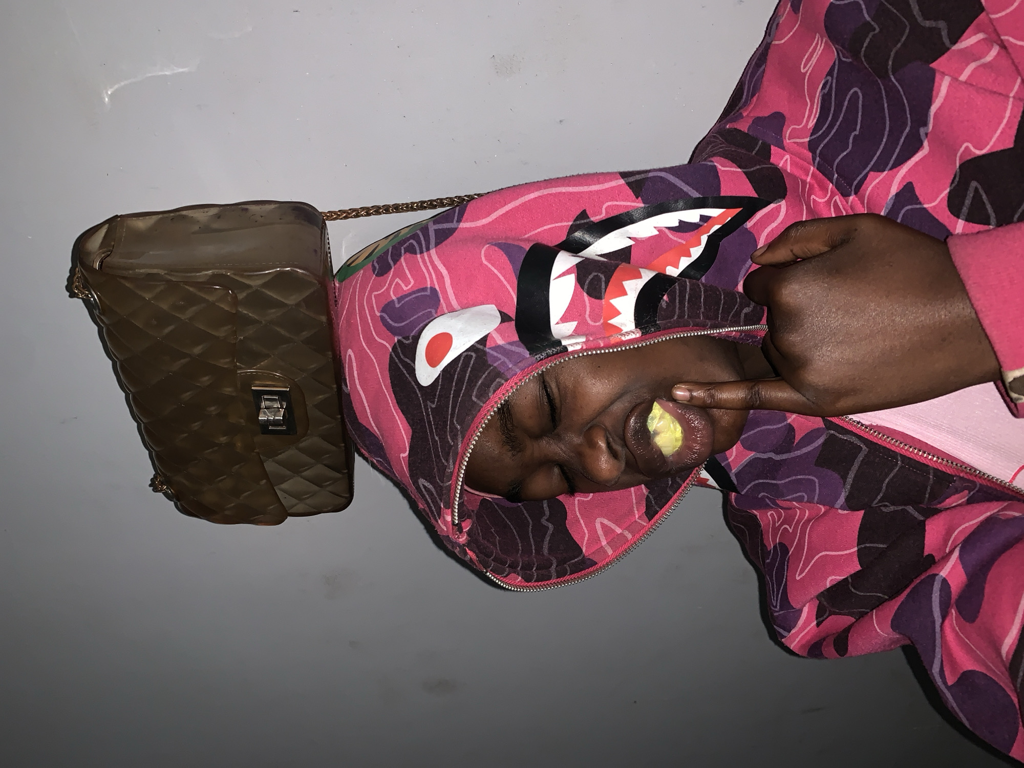 A person wearing a pink and purple camouflage hoodie is eating something yellow while posing for a photo. The person is wearing a clear brown quilted handbag with a gold chain strap over the shoulder. The background is a plain gray wall. The location is Rafin Par, Nigeria.