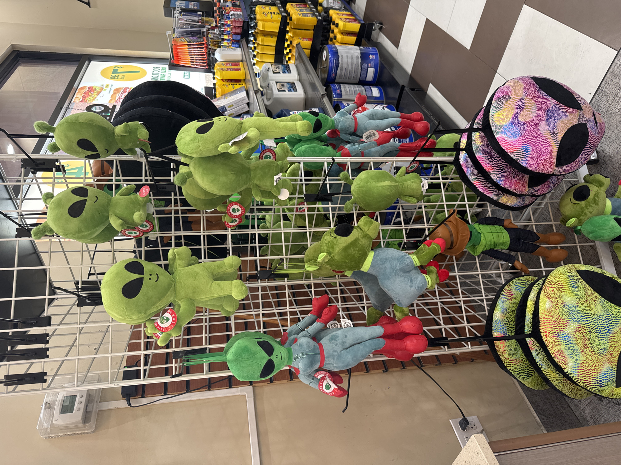 The image shows a retail display of stuffed alien toys, some hanging from a wire rack and others stacked on a lower shelf. A few of the alien toys are green with large black eyes, while others have different colored, patterned heads. In the background, shelves are stocked with various products, including yellow bottles and blue containers, suggesting a store environment. There is a thermostat mounted on the wall to the left of the display. The flooring appears to be tiled in a checkered pattern. The scene is likely a store in Baker, United States of America, possibly a gas station or convenience store given the products on the shelves.