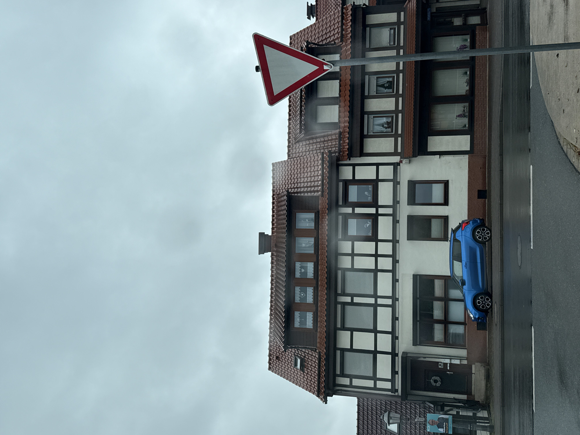 This is a medium shot, taken from a street-level perspective, of a residential building and a yield sign on a cloudy day in Bad Sachsa, Germany. The building, a two-story structure with a red tile roof, features traditional German half-timbered architecture with white stucco walls and dark brown beams. Several windows are visible, some with white blinds and others decorated with lace curtains. A blue compact car is parked on the wet street in front of the building. To the right of the building, a silver yield sign on a metal pole stands prominently. The street in the foreground is wet, indicating recent rain. A lamppost and a political poster are visible to the left of the building. The sky is overcast and light gray.