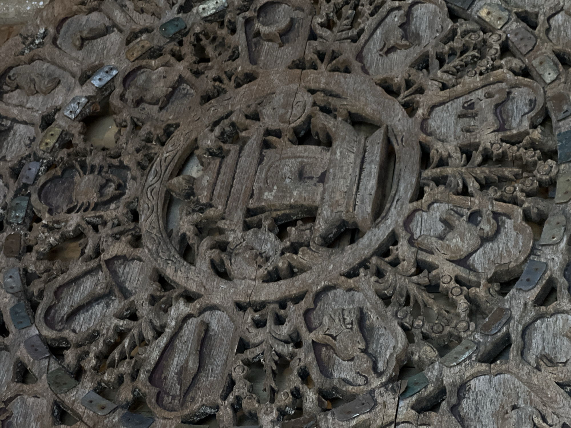 This image presents a detailed close-up of an aged and intricately carved wooden surface, possibly a panel, door, or piece of furniture. The wood displays a weathered, greyish-brown texture with visible grain, cracks, and signs of erosion, suggesting considerable age.
At the center of the carving is a prominent circular motif enclosing what appears to be a stylized architectural structure, possibly a temple, gate, or building with distinct pillars and a roof-like element. Surrounding this central design is a complex array of smaller, interconnected carvings featuring various organic, floral, and symbolic patterns. Among these, several distinct animal figures can be discerned, such as a creature resembling an elephant's head in the upper-left section and another with what looks like horns or antlers in the lower-right area.
Numerous small, rectangular metal inserts are interspersed across the carved surface. These flat pieces, which vary in color from silver-toned to brassy yellow and greenish (indicating oxidation), each typically feature two small holes, suggesting they were once affixed with fasteners, possibly serving as decorative elements or repairs. The overall craftsmanship is highly detailed, showcasing a complex interplay of deep relief and delicate lines. The scene is static, depicting only the carved object. There is no visible text.