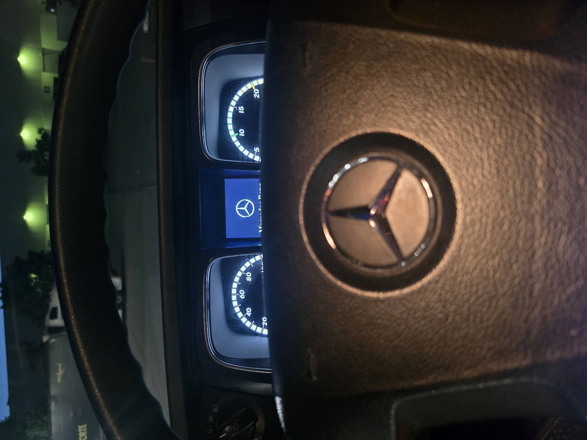 The image shows the interior of a Mercedes-Benz truck at night. The perspective is from the driver's seat, looking forward. The steering wheel, with the Mercedes-Benz logo prominently displayed, is in the foreground. Behind the steering wheel are two digital instrument clusters. The left cluster displays a speedometer with markings from 0 to 100, indicating a current speed of approximately 20. The right cluster shows a gauge with markings from 0 to 20, with a needle pointing between 15 and 20, possibly representing engine RPM or fuel level. A blue screen on the left displays the Mercedes-Benz logo. Outside the vehicle, dimly lit by green lights, are buildings and trees, suggesting an industrial or commercial area. The scene is captured in low light conditions, typical of nighttime. The location is identified as Zeppelinheim, Germany.