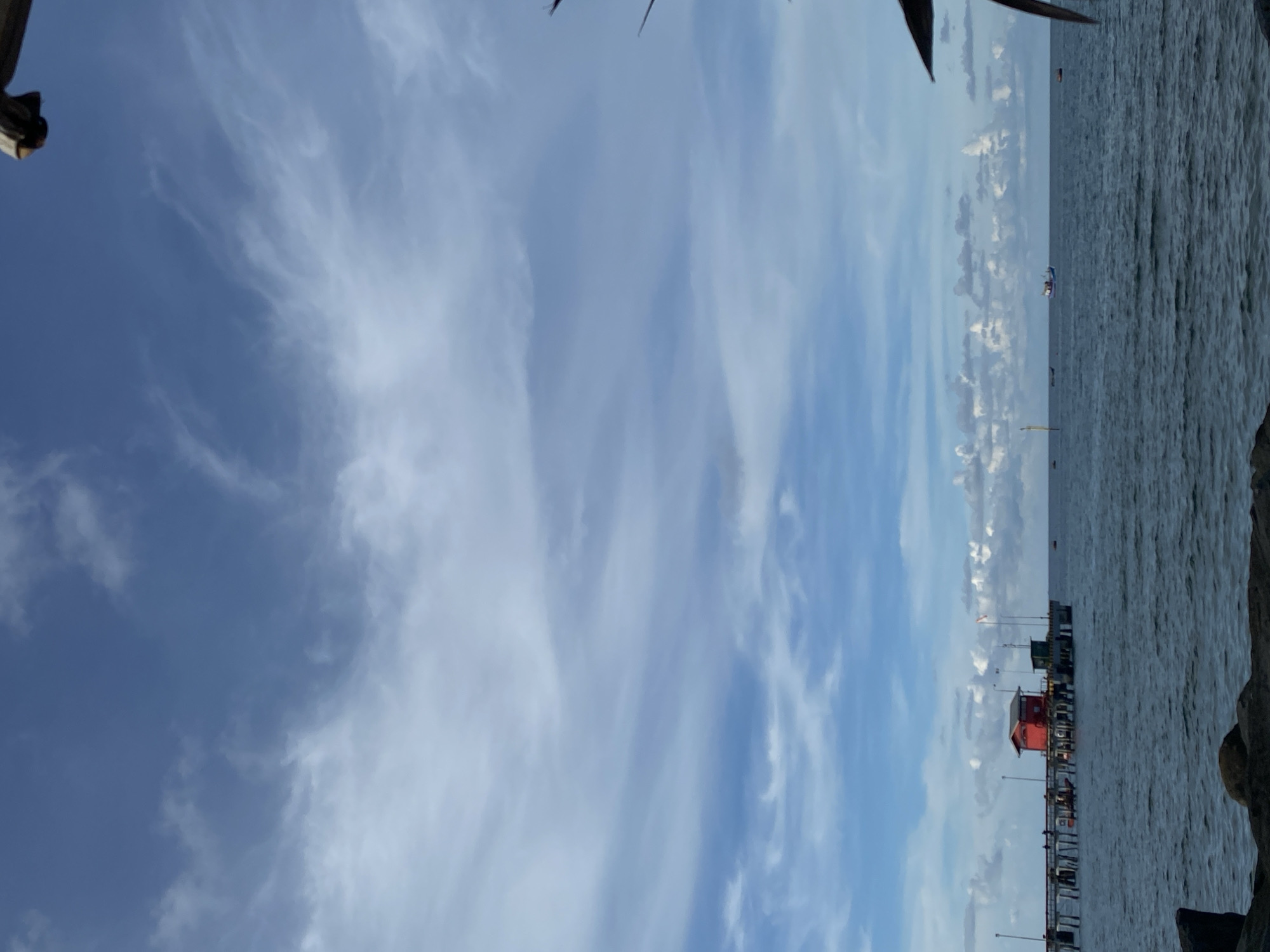 The image shows a vertical view of a waterfront scene. On the right side, there is water, and a pier or dock extends out into the water. Several small structures are on the pier, including a red building. In the distance, there is at least one boat or ship visible on the water. The sky is predominantly blue with wispy white clouds. The location is likely Lhokseumawe, Indonesia, due to the style of the structures and the landscape. There is a sense of calm and openness to the scene.