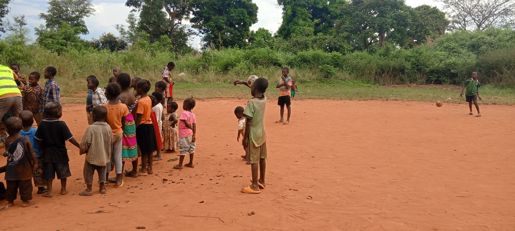 In Ngalula, Democratic Republic of the Congo, a group of approximately 25 children and one adult stand on a dirt field bordered by green foliage and trees. The children wear a variety of casual clothing and sandals, while the adult wears a yellow vest. A boy stands apart from the group, dribbling a soccer ball. Another boy holds a soccer ball up as if getting ready to throw it.