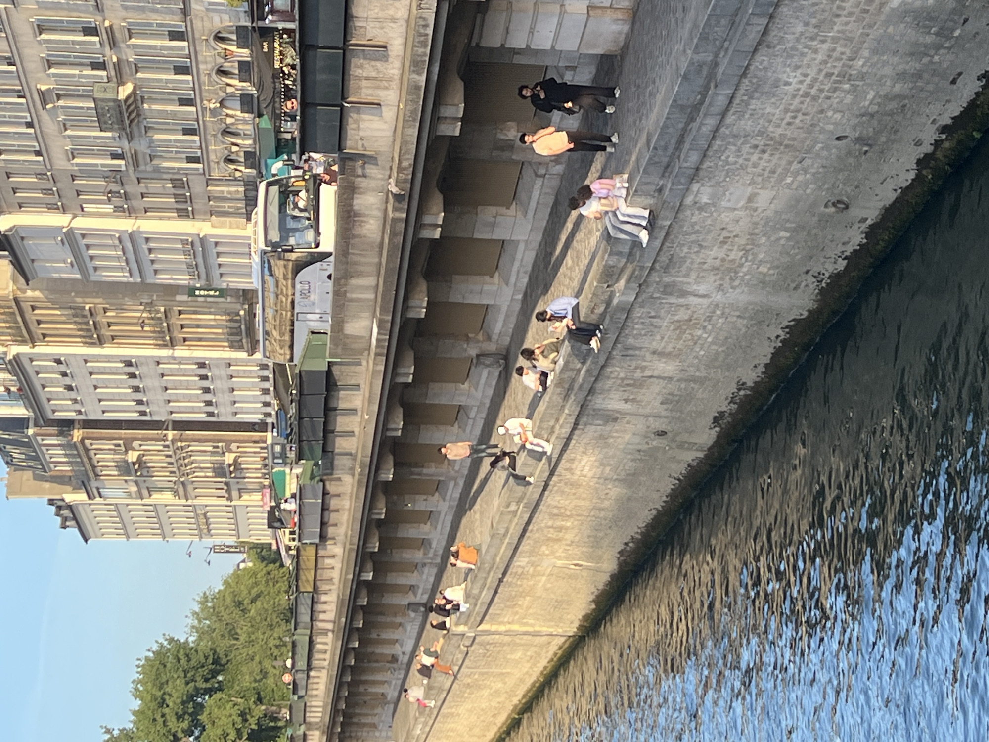 The image depicts a sunny afternoon scene along the Seine River in Paris, France. A stone embankment lines the river, with a pathway on top where numerous individuals are seated and standing. The pathway is bordered by a building facade with multiple stories, featuring numerous windows and architectural details characteristic of Parisian architecture.
On the pathway, groups of people are casually interacting. Some are sitting on the edge of the embankment, facing the river, while others are gathered in small groups on the pathway itself. The attire of the individuals suggests a warm weather day, with many wearing casual clothing such as t-shirts, shorts, and dresses. The river is visible in the foreground, reflecting the sunlight and the surrounding buildings.
Further back, a white tour bus with "APOLLO" branding is parked, indicating tourist activity in the area. Several awnings and small structures are visible along the building facade, suggesting shops or cafes. The overall atmosphere is relaxed, with people enjoying the riverside ambiance. The lighting suggests it is late afternoon, with the sun casting long shadows.