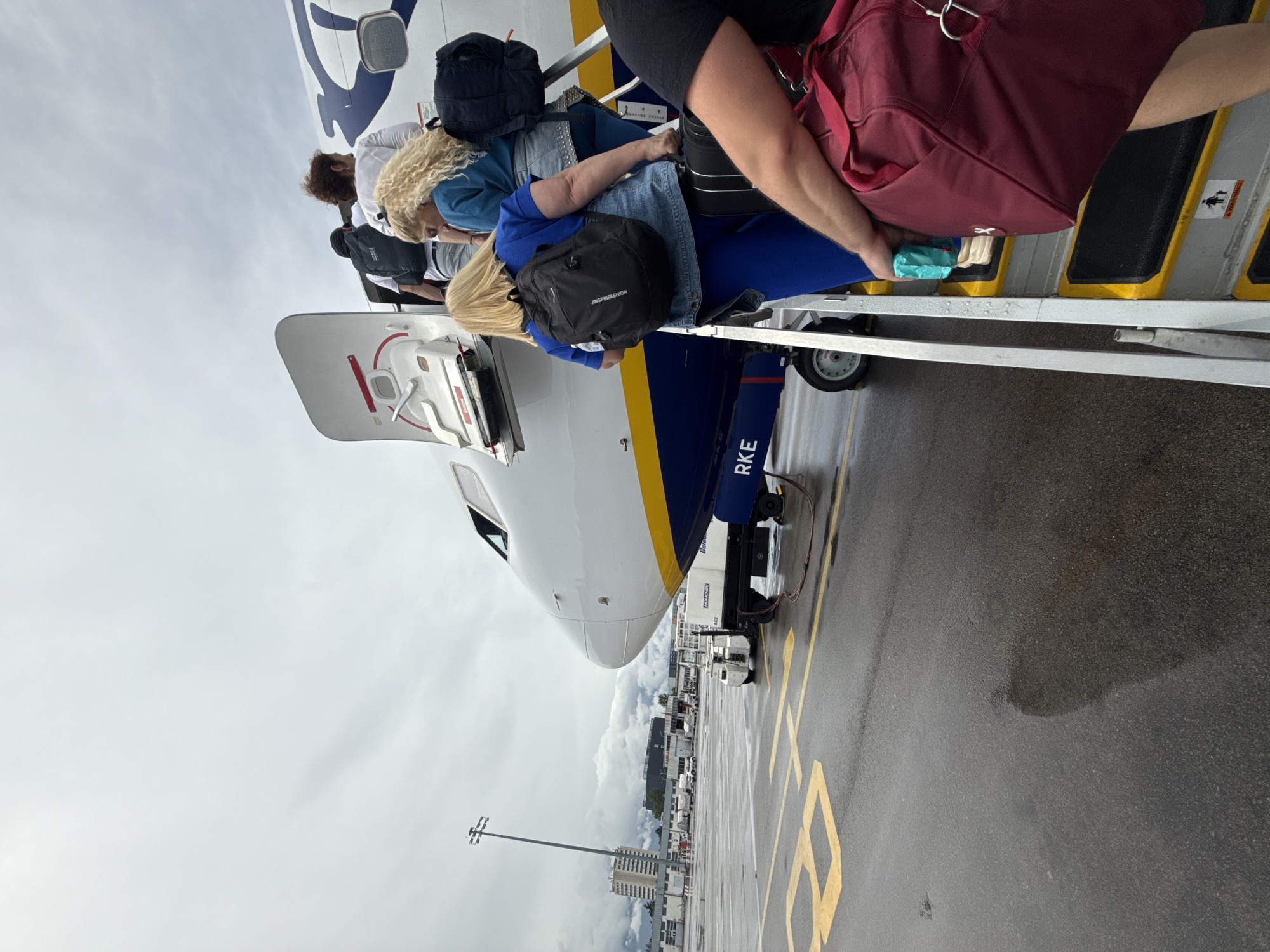 An overhead view captures passengers boarding an aircraft on an airport tarmac. The image is taken from a slightly elevated position, looking down the length of the plane. Several individuals, mostly women, are seen climbing mobile stairs into the aircraft, their backs to the camera. They are carrying various personal bags, including backpacks and duffel bags.
The aircraft itself features a white fuselage with a prominent blue stripe along its side, characteristic of certain airlines like Ryanair. Part of what appears to be an engine or rear fuselage section bears the partial lettering "RKE." The nose of the plane is visible further down the tarmac.
The weather appears overcast, with a light gray sky dominating the upper left portion of the image. The asphalt tarmac is wet, suggesting recent rain. Yellow painted lines and markings, including a partially visible "1B," are on the ground. In the background, beyond the aircraft, airport buildings, and a tall light pole are discernible under the cloudy sky, placing the scene at an airport in Sofia, Bulgaria.
Visible text includes "JINGPINFASHION" on a black backpack carried by one of the passengers. A "WARNING" label with a graphic of a person falling is present on the boarding stairs. The activity depicts a standard boarding procedure for an airplane, with passengers ascending the stairs to enter the cabin.