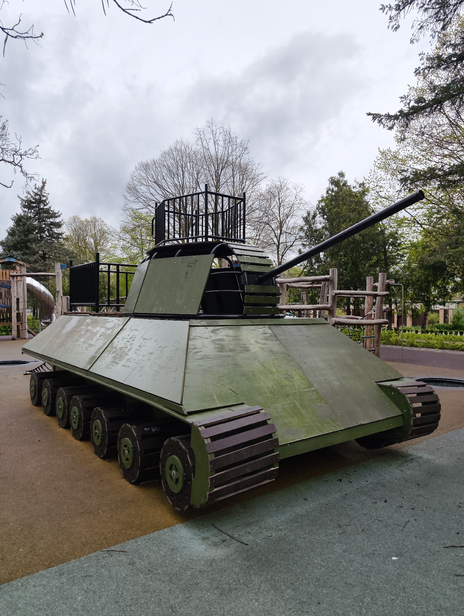 The image shows a wooden tank playground structure located in a park. The tank is painted green and has a black barrel. It has several wheels on each side and wooden tracks. There is a black metal railing on top of the tank. In the background, there are trees and other playground structures. The picture was taken in Krasnodar, Russia.