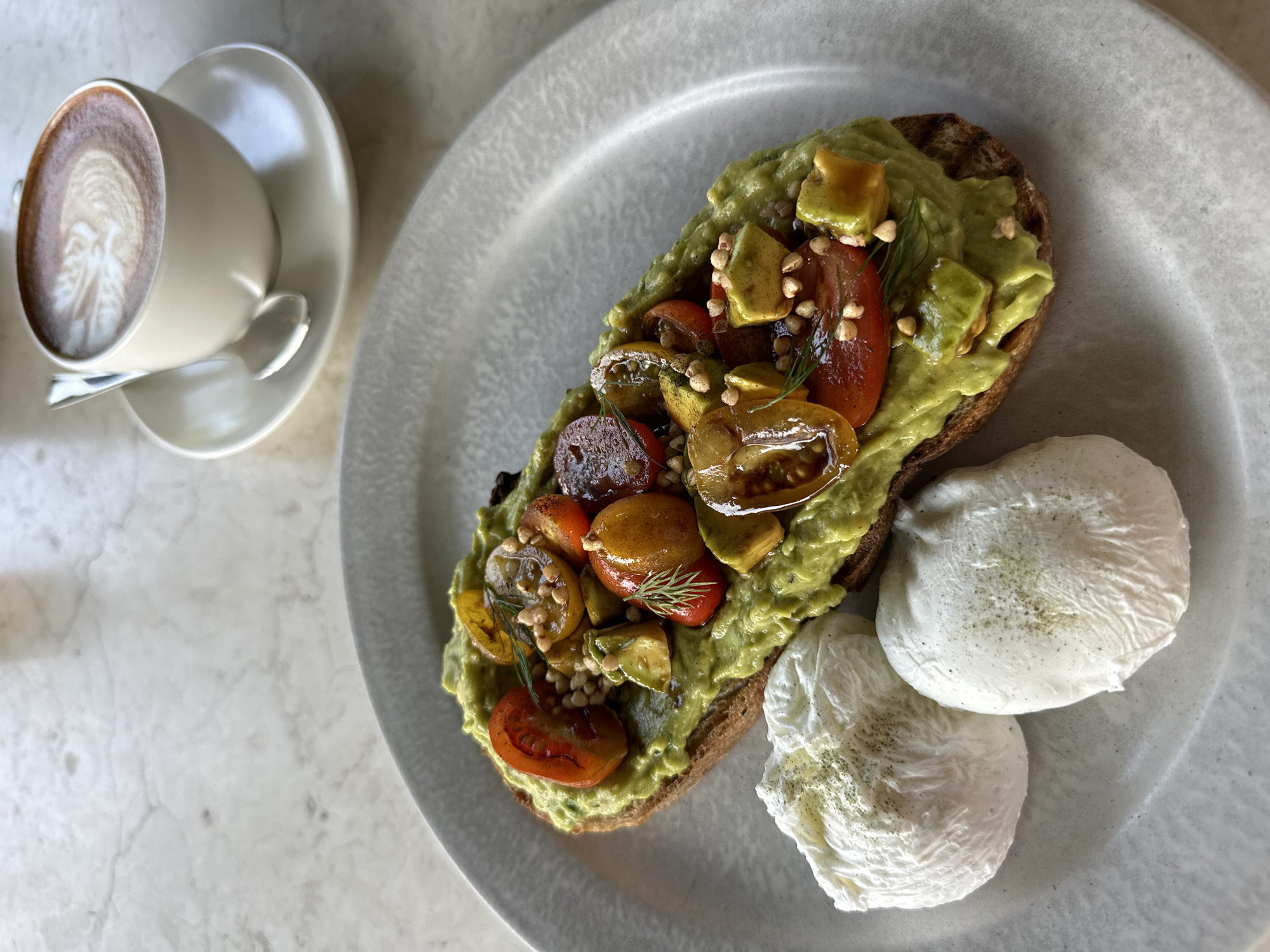The image shows a plate of avocado toast with two poached eggs, next to a cup of coffee. The toast is topped with mashed avocado, sauteed tomatoes, and small seeds. The coffee is a latte with latte art. The scene appears to be in a cafe or restaurant. The location is Unknown, Unknown.