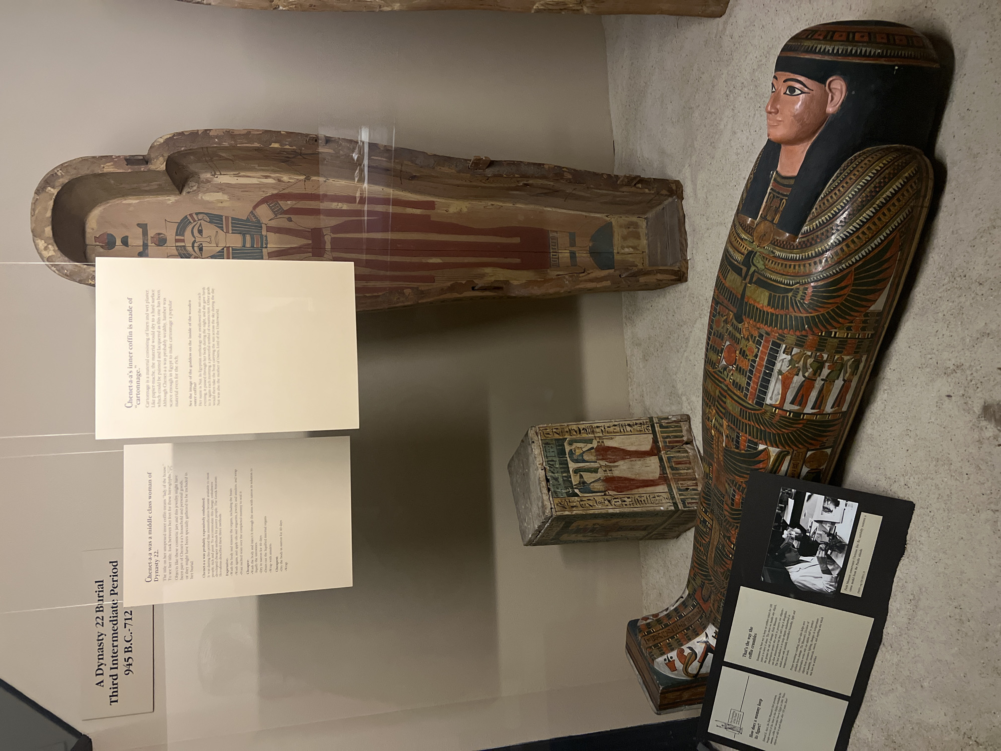 This image shows a display of ancient Egyptian artifacts, likely in a museum setting. The display includes a sarcophagus with painted figures, an inner coffin made of cartonnage, a canopic jar (likely for storing organs), and various informational plaques. The informational plaques provide details about the artifacts, including the name of the individual ("Chenet-a-a"), the period (Dynasty 22, Third Intermediate Period, 945 B.C.-712), and the materials used in the construction of the coffin. Another plaque shows a black and white photo from the conservation effort of the artifacts. The artifacts are under glass. Given the presence of a display and informational plaques, the location is most likely the Art Institute of Chicago in Chicago, United States of America.