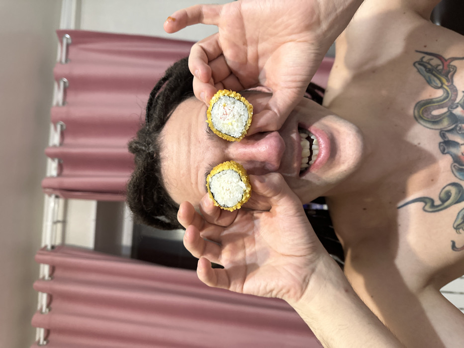 A man with dreadlocks holds two pieces of sushi over his eyes. He is shirtless and has a tattoo on his left shoulder. He is smiling wide showing his teeth. A pink curtain or window shade is in the background.