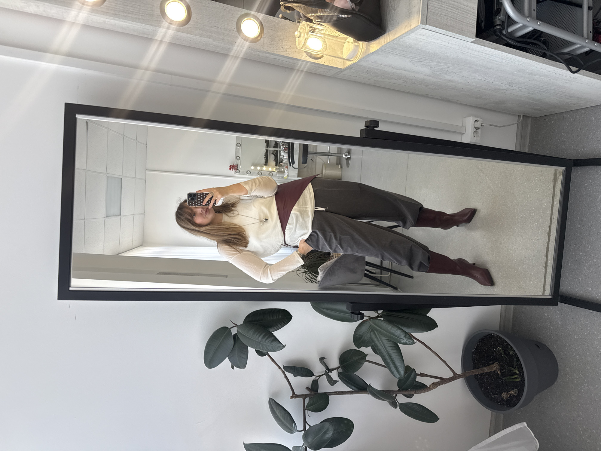 A full-body shot depicts a female subject taking a mirror selfie within what appears to be a beauty or makeup studio in Izhevsk, Russia.
**Subject:**
A female with light brown hair, reaching past her shoulders, stands facing the mirror directly. She is wearing a long-sleeved, translucent white top, dark grey or charcoal wide-leg pants with a white drawstring at the waist, and pointed-toe, high-heeled burgundy boots. A wide burgundy sash or belt is wrapped diagonally around her waist over her top. A silver-colored necklace with a prominent pendant is visible. Her left hand is holding a black smartphone with a white polka-dot case, aimed at the mirror to capture the image. Her right hand is in the pocket of her pants. She has light makeup visible on her face.
**Environment (reflected in mirror):**
The reflection reveals a well-lit, white-walled room with a suspended acoustic panel ceiling featuring rectangular light fixtures. To the right of the subject, a vanity mirror with circular, exposed light bulbs around its frame is visible, alongside a light-colored wooden or laminate counter holding various beauty items, including what appears to be a clear glass container with white pads and a dark bag. A dark-colored, backless stool is situated in front of the vanity. Further back, another grey upholstered chair is visible, along with other studio equipment or furniture, including a rolling cart with shelving. The overall impression is a clean, professional interior setting.
**Immediate Surroundings (outside reflection):**
To the left of the mirror, a potted Ficus elastica (rubber plant) with large, dark green leaves stands on the light grey speckled floor. The wall behind the plant is plain white. Below the vanity counter and near the floor on the right side of the mirror, a white power outlet with two sockets is visible, with a white cord plugged into one. The floor throughout is a light grey speckled material. The mirror itself has a thin black frame and is freestanding on a black stand.