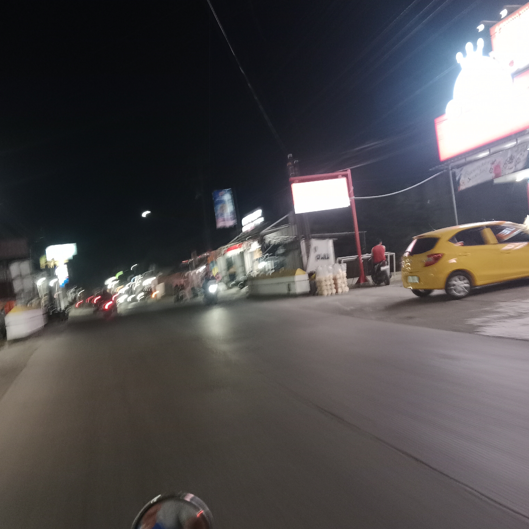 The image captures a nighttime scene on a road in Gubug, Indonesia. A yellow car is parked on the right side of the road, facing away from the camera. To the left of the car, a person in a red shirt is seated on a motorcycle. Further down the road, multiple vehicles are visible, their headlights and taillights creating streaks of light due to the motion blur. Buildings and streetlights line the sides of the road, with illuminated signs indicating commercial activity. The road surface appears wet, reflecting the surrounding lights. The overall impression is of a busy street at night.