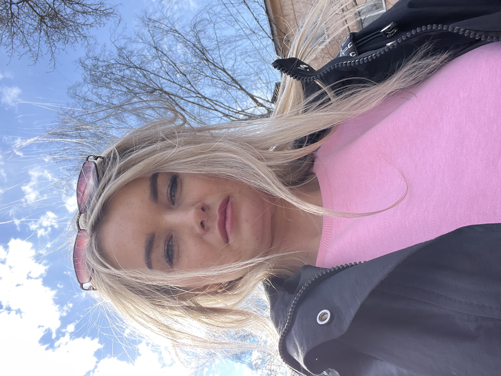 The image is a rotated close-up selfie featuring a young woman outdoors in Chaikovsky, Russia. She has long, windswept blonde hair, fair skin, and light-colored eyes, with light makeup visible on her eyelids and lips. Sunglasses are perched on top of her head. Her expression is neutral, with a slight pout.
She is wearing a ribbed, light pink long-sleeved top or sweater, mostly covered by a black jacket. The jacket features a prominent silver zipper and at least one silver eyelet or snap.
The background consists of a bright blue sky with scattered white clouds, visible above and to the left of the woman. Silhouetted against the sky are numerous bare tree branches, suggesting a season like late winter or early spring. The bright sunlight indicates it is daytime, likely mid-morning or afternoon. No discernible text is visible within the frame. The photograph appears to have been taken from a low angle, looking upwards towards the subject against the sky.